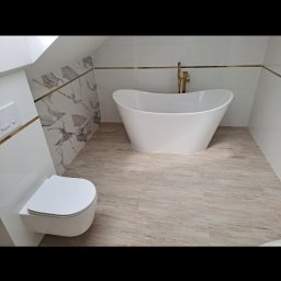 Marcin Głowiak - Nowoczesna łazienka na poddaszu z wolnostojącą wanną, podwieszaną toaletą i marmurowymi akcentami. Złote detale armatury i listew podkreślają elegancję wnętrza.