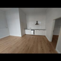 Marcin Głowiak - Jasne, przestronne wnętrze po remoncie: drewniana podłoga, białe ściany i zabudowa kuchenna z okapem. Widok na otwarte drzwi do kolejnego pomieszczenia.