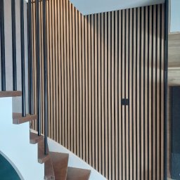 Marcin Głowiak - Nowoczesne schody z drewnianymi stopniami i czarną balustradą na tle ściany z paneli dekoracyjnych w odcieniach beżu i czerni. Minimalistyczny design.