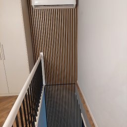 Marcin Głowiak - Nowoczesne wykończenie ściany z drewnianych lameli w pionowym układzie, z zamontowanym klimatyzatorem. Widok na schody z czarną balustradą i białą poręczą.
