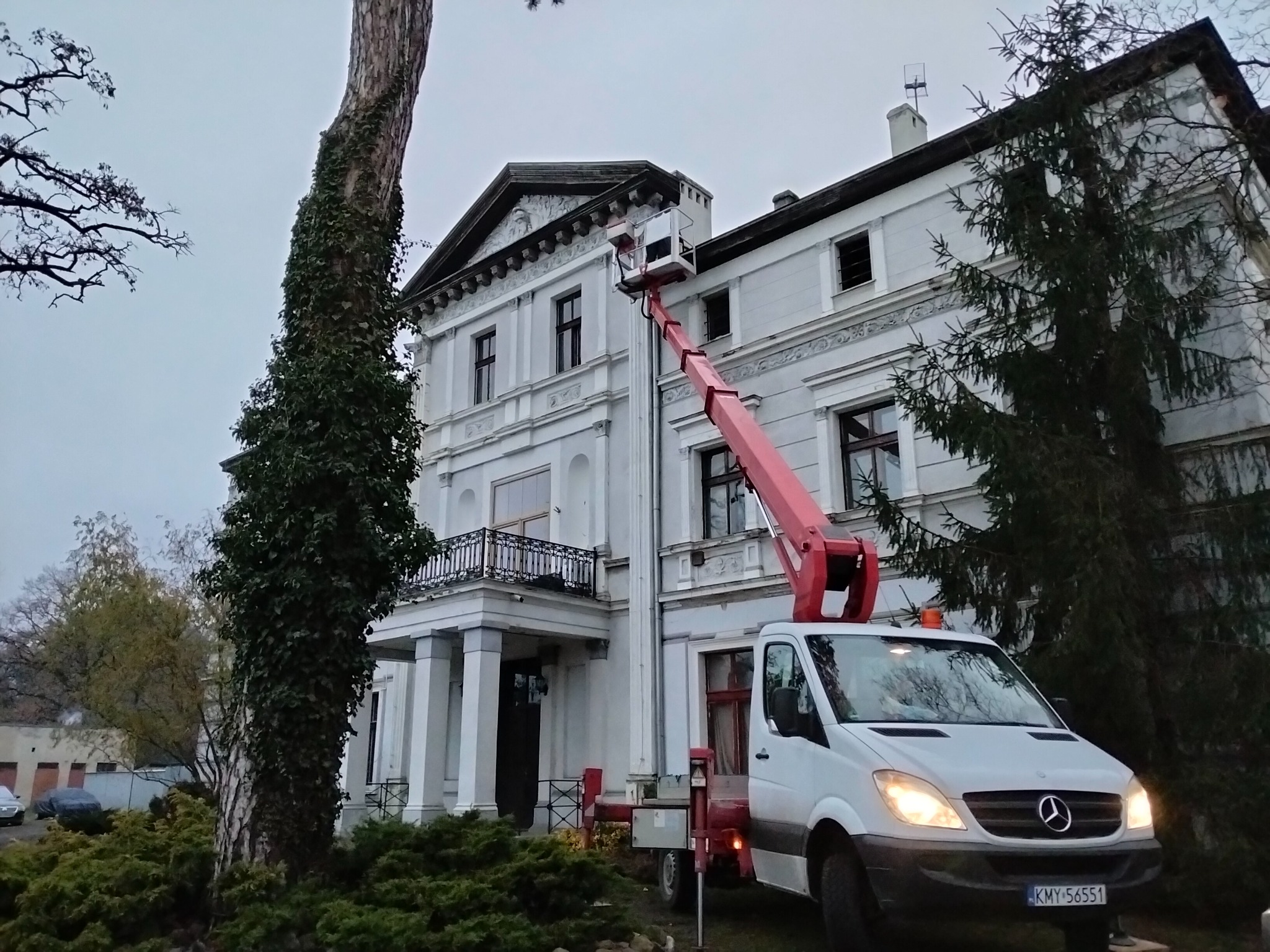 Prace wysokościowe przy zabytkowym, dwupiętrowym budynku z kolumnami. Czerwony podnośnik koszowy na białym samochodzie dostawczym sięga do dachu.