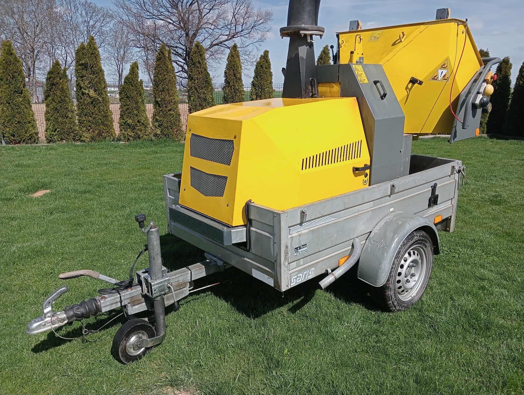 Żółto-szara rębarka do gałęzi na przyczepie, stojąca na trawie. W tle rząd tuj i drzewa. Sprzęt gotowy do pracy w ogrodzie.