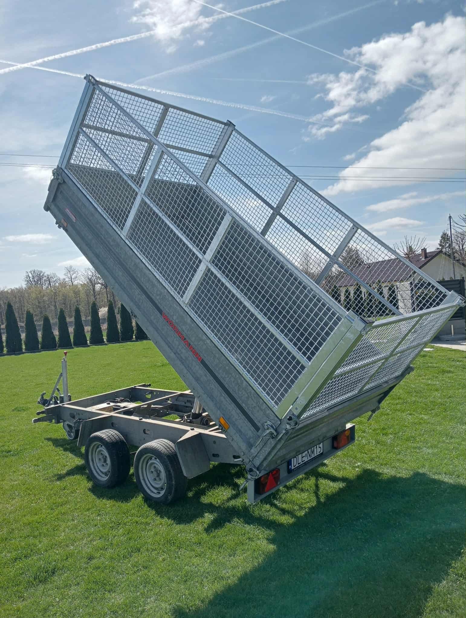 Przyczepa trójosiowa z podniesioną skrzynią ładunkową z siatki, zaparkowana na trawniku w słoneczny dzień. Widoczne tablice rejestracyjne.