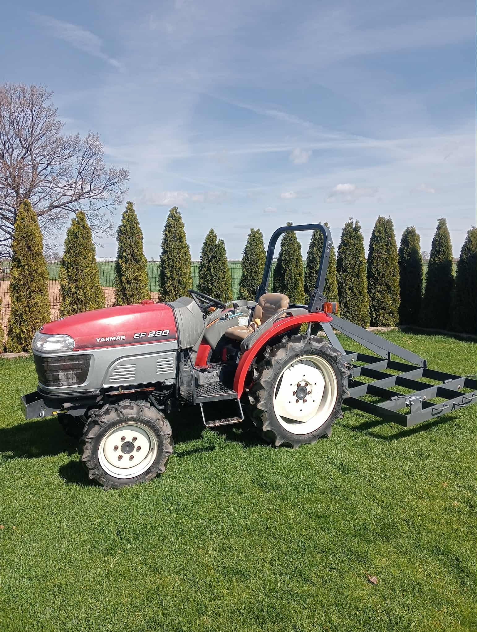 Czerwono-szary traktor Yanmar EF 220 z zamontowanym sprzętem na trawniku, w tle rząd tuj i błękitne niebo. Sprzęt do pielęgnacji trawnika.