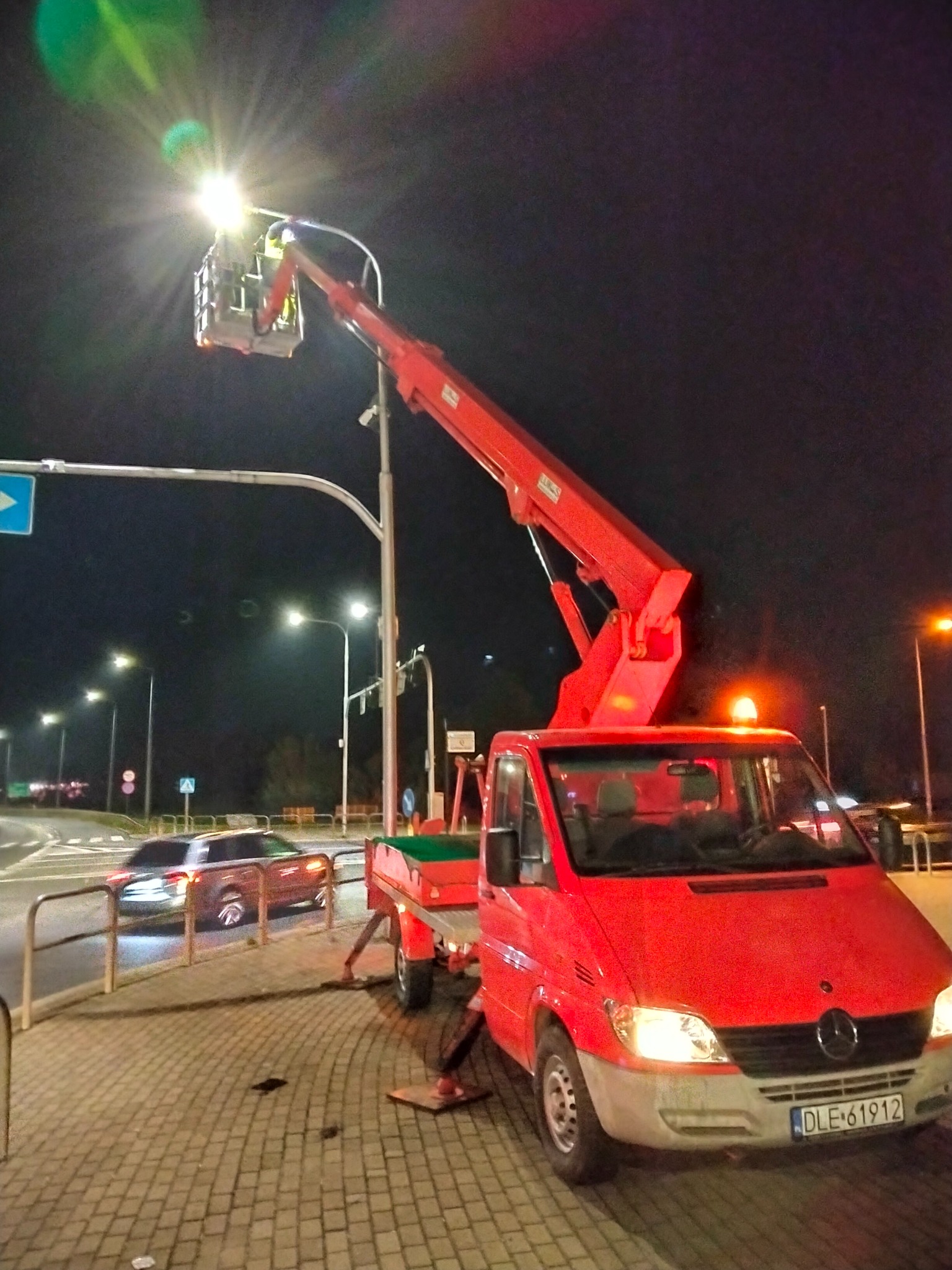 Czerwony podnośnik koszowy Mercedes Sprinter oświetla nocną ulicę podczas pracy, widoczny pracownik w koszu i zaparkowany samochód osobowy w tle.