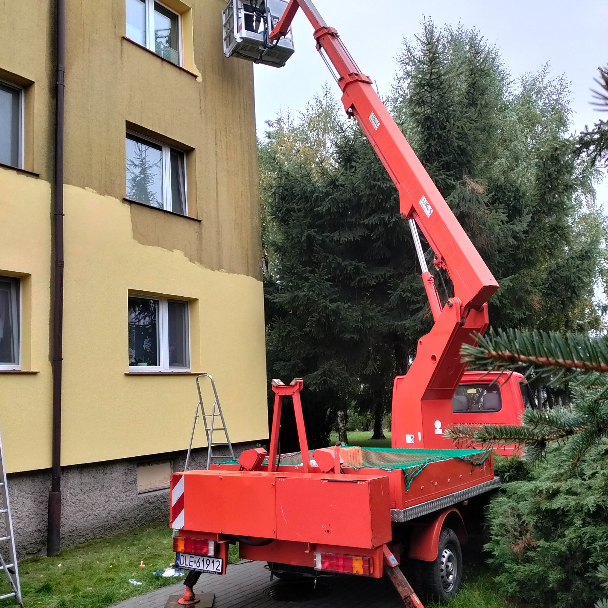 Czerwony podnośnik koszowy przy żółtej elewacji budynku w trakcie malowania. Widoczny fragment pomalowanej i niepomalowanej ściany. Na dole drabina.