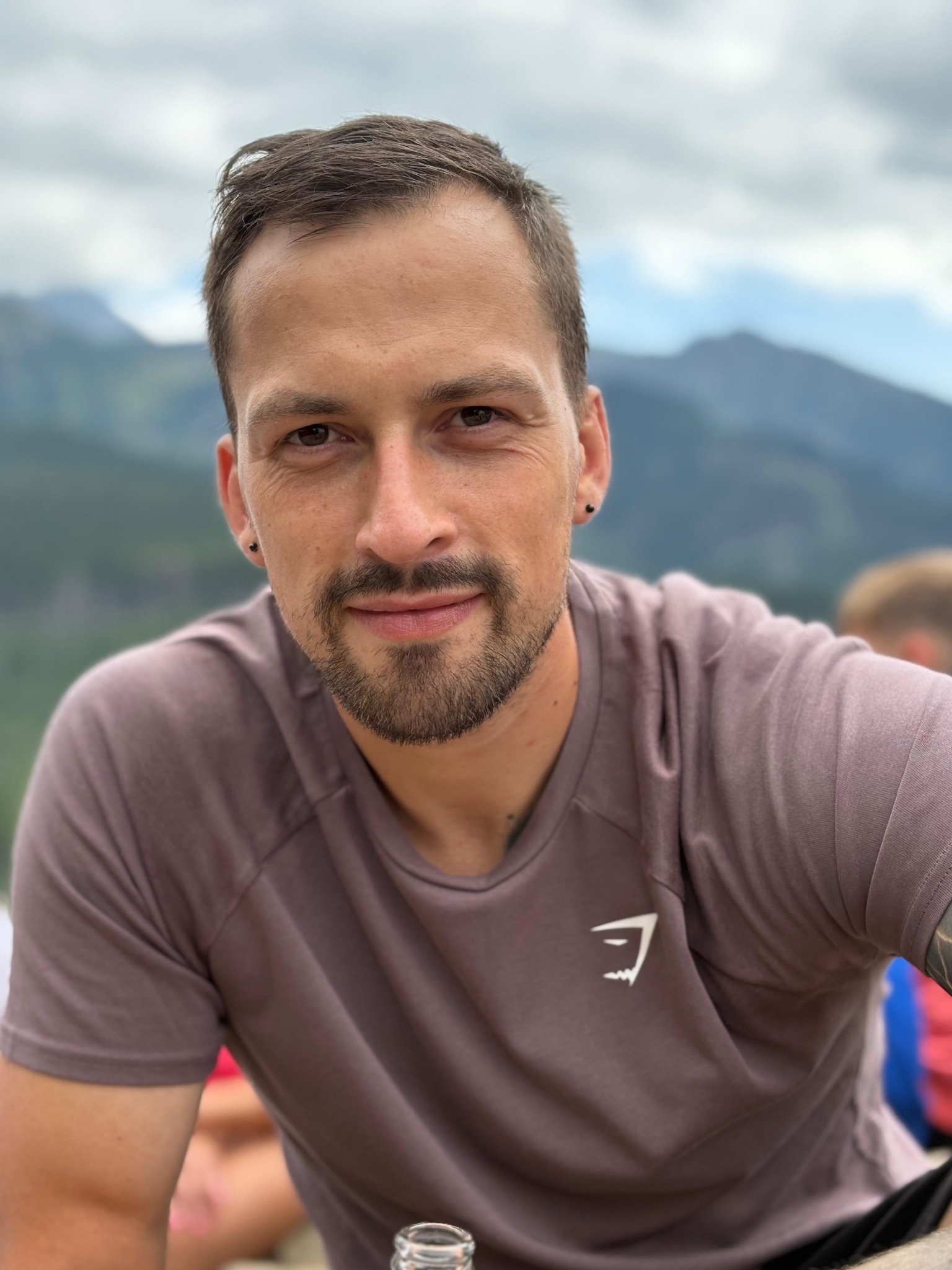 Portret mężczyzny z brodą na tle gór. Ubrany w koszulkę z logo. Widoczne kolczyki w uszach. Uśmiechnięty, patrzy w obiektyw. Naturalne światło.