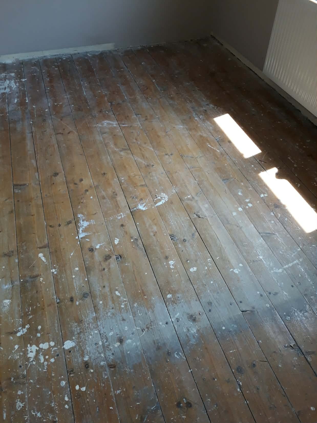 Drewniana podłoga z widocznymi śladami białej farby i smugami światła słonecznego w rogu pokoju, ukazująca stan przed renowacją.