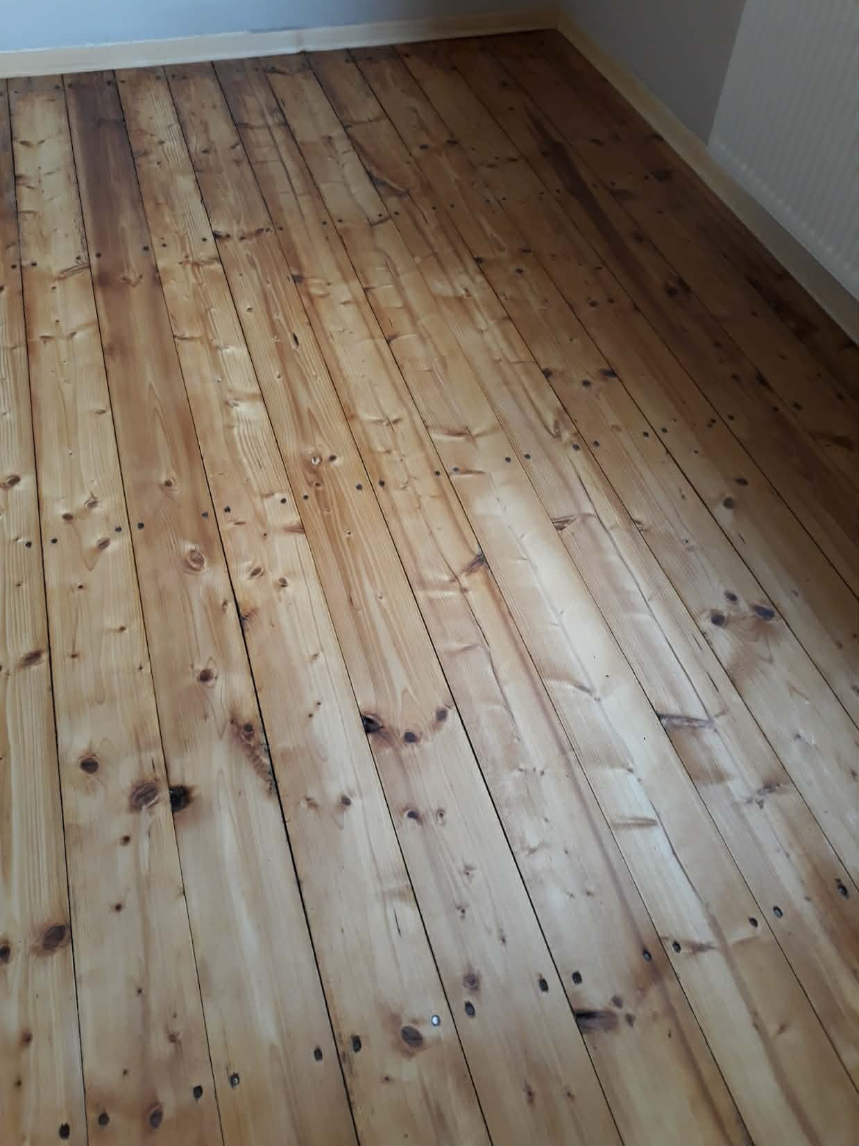 Jasna, drewniana podłoga z widocznymi śladami po gwoździach i naturalnym usłojeniem, ułożona wzdłuż pomieszczenia, z listwami przypodłogowymi w tle.