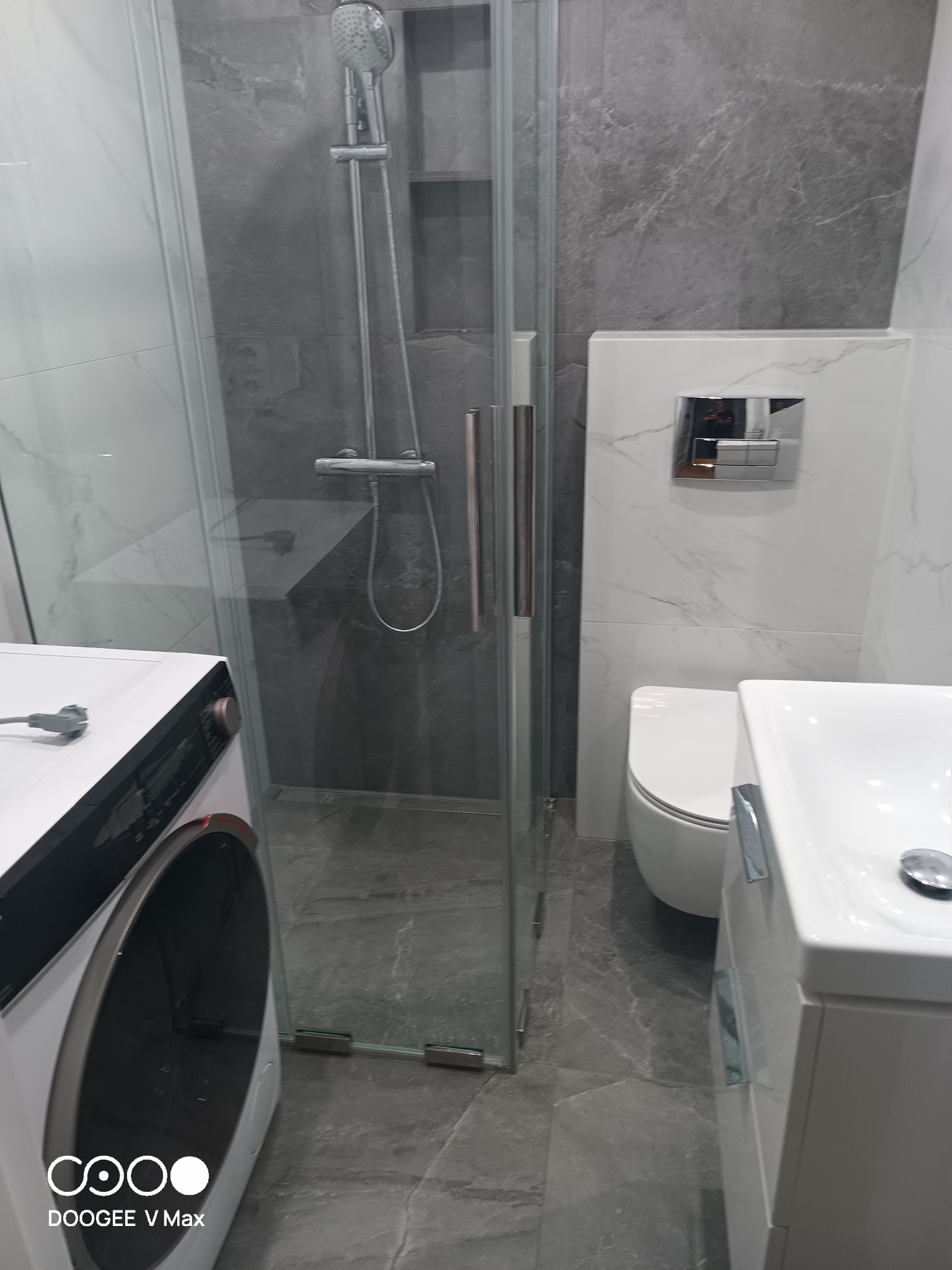 Nowoczesna łazienka z kabiną prysznicową typu walk-in, umywalką na szafce i pralką. Szare płytki imitujące marmur na ścianach i podłodze. Minimalistyczny design.