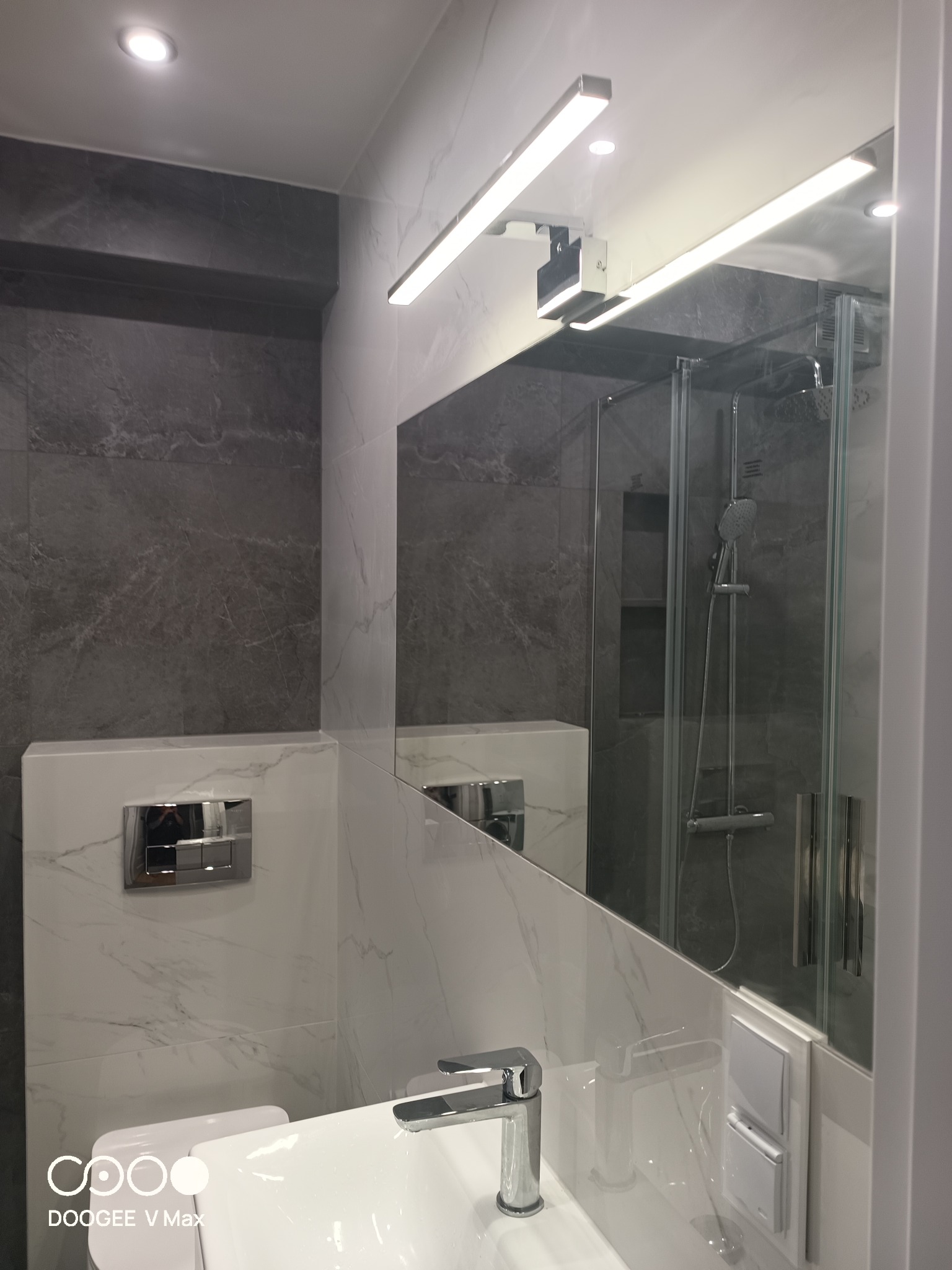 Nowoczesna łazienka z szarymi marmurowymi płytkami, białym umywalką i kabiną prysznicową odbijającą się w lustrze. Oświetlenie LED nad lustrem.