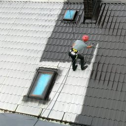 Alame - Pracownik w kasku, zabezpieczony linami, maluje dachówkę na dachu z oknami dachowymi, używając pistoletu natryskowego. Część dachu jest już pomalowana na ciemny kolor.