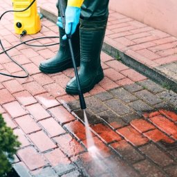 Alame - Czyszczenie kostki brukowej myjką ciśnieniową. Widoczne różnice w kolorze kostki przed i po czyszczeniu. Pracownik w gumowych butach i rękawicach ochronnych.