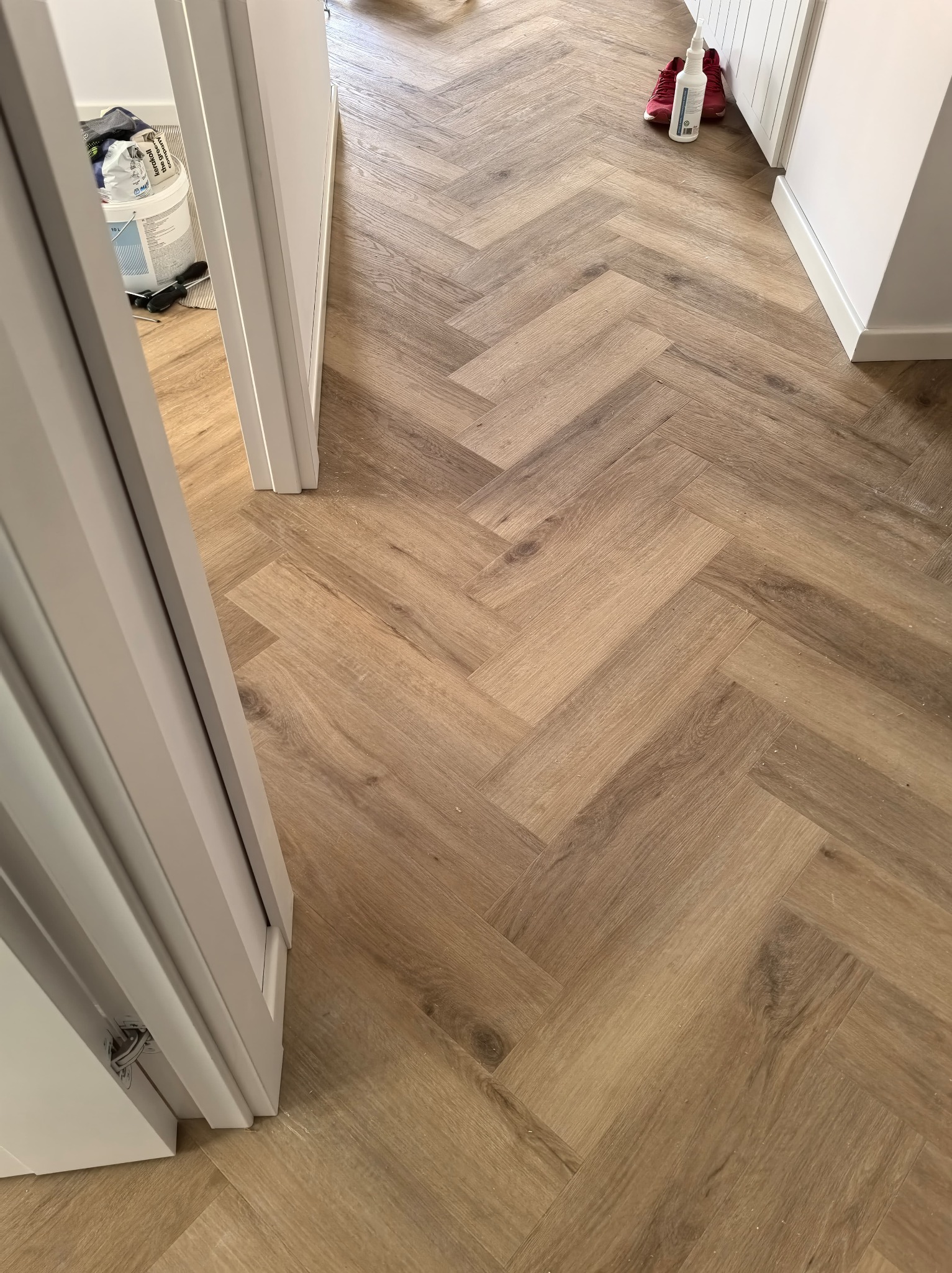 Ułożona podłoga z paneli w jodełkę, widok z perspektywy wejścia do pomieszczenia. W tle widoczne narzędzia i akcesoria malarskie.