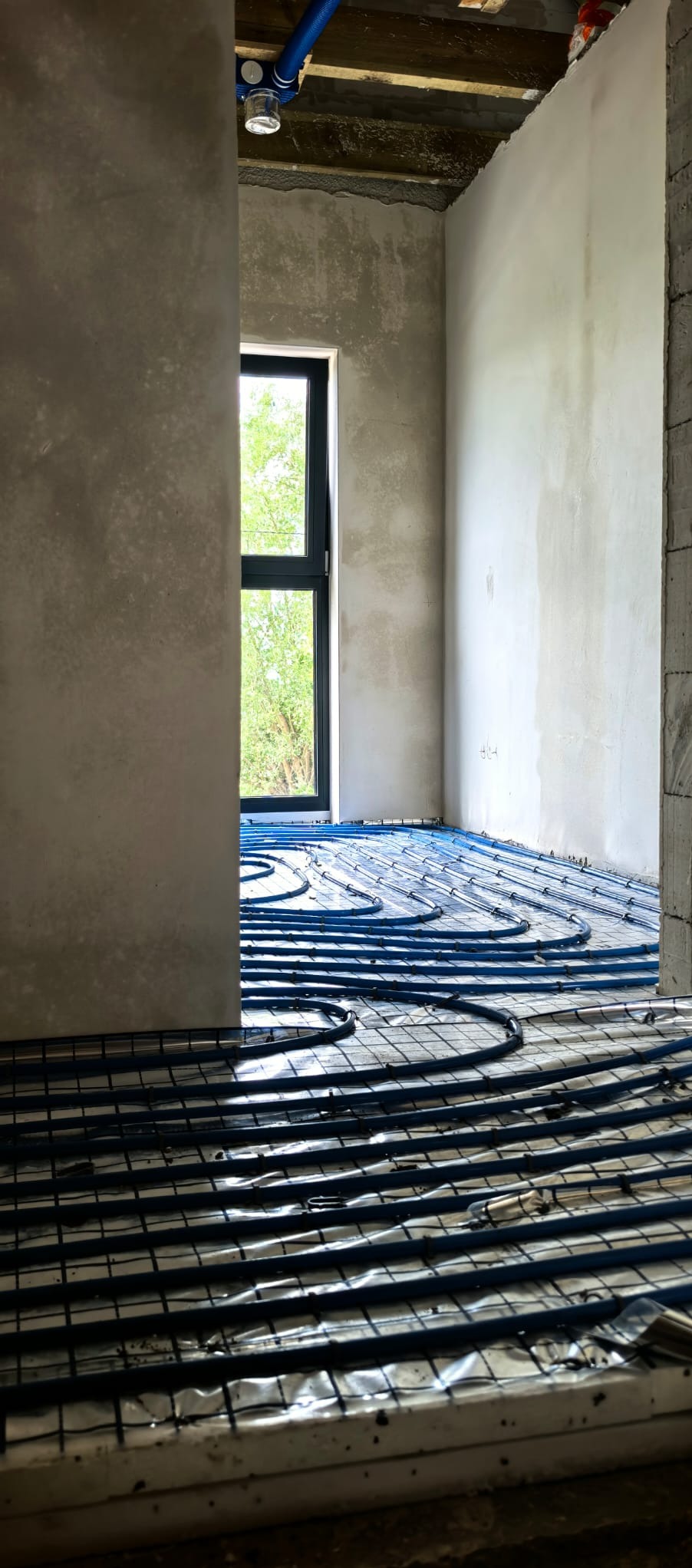 Niebieskie rury ogrzewania podłogowego ułożone na folii w surowym wnętrzu z oknem. Widoczne ściany w szarym tynku i fragment drewnianego stropu z instalacją.