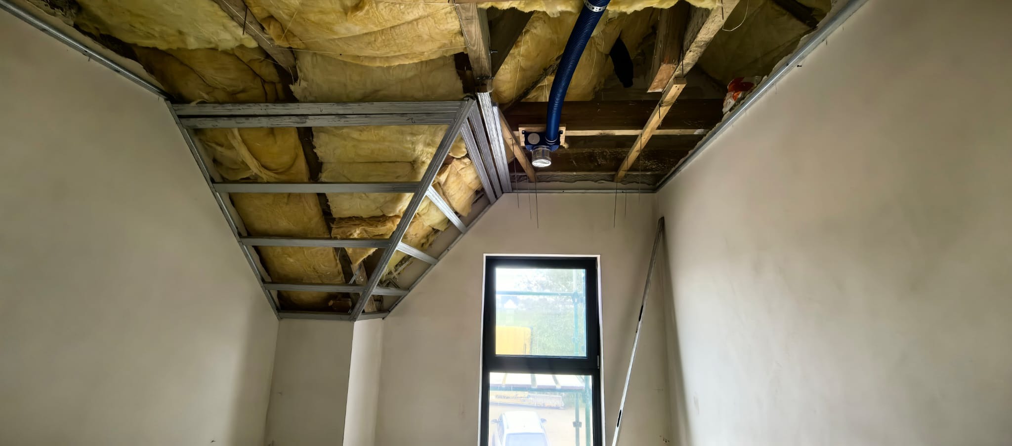 Skośny sufit z izolacją z wełny mineralnej i metalowym stelażem pod płyty gipsowe, widok na okno i częściowo wykończone ściany w jasnym pomieszczeniu.