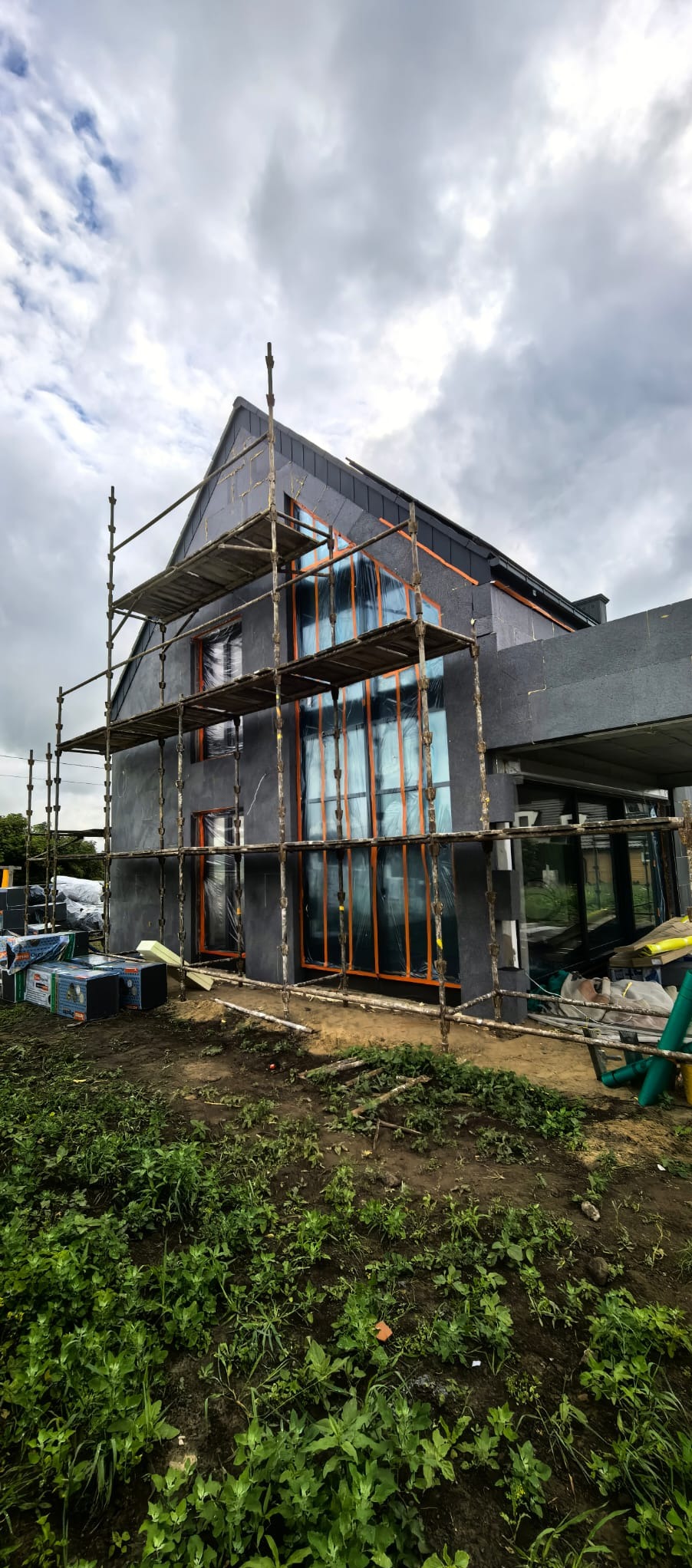 Dom w trakcie prac wykończeniowych z rusztowaniem. Okna zabezpieczone folią, elewacja w szarym kolorze. Widoczne materiały budowlane i roślinność.