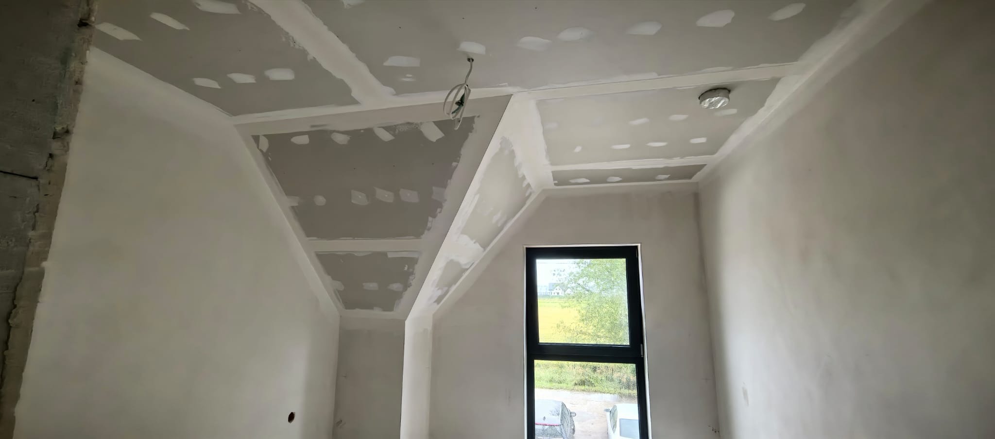 Skośny pokój po montażu płyt karton-gips na suficie i ścianach, widoczne łączenia płyt przygotowane do szpachlowania. Okno w czarnej ramie.