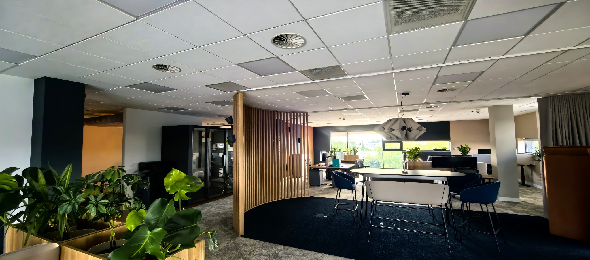 Nowoczesne biuro po remoncie: jasne wnętrze z roślinami, designerskim oświetleniem i meblami. Widok na przestrzeń open space z elementami drewna i ciemnymi akcentami.