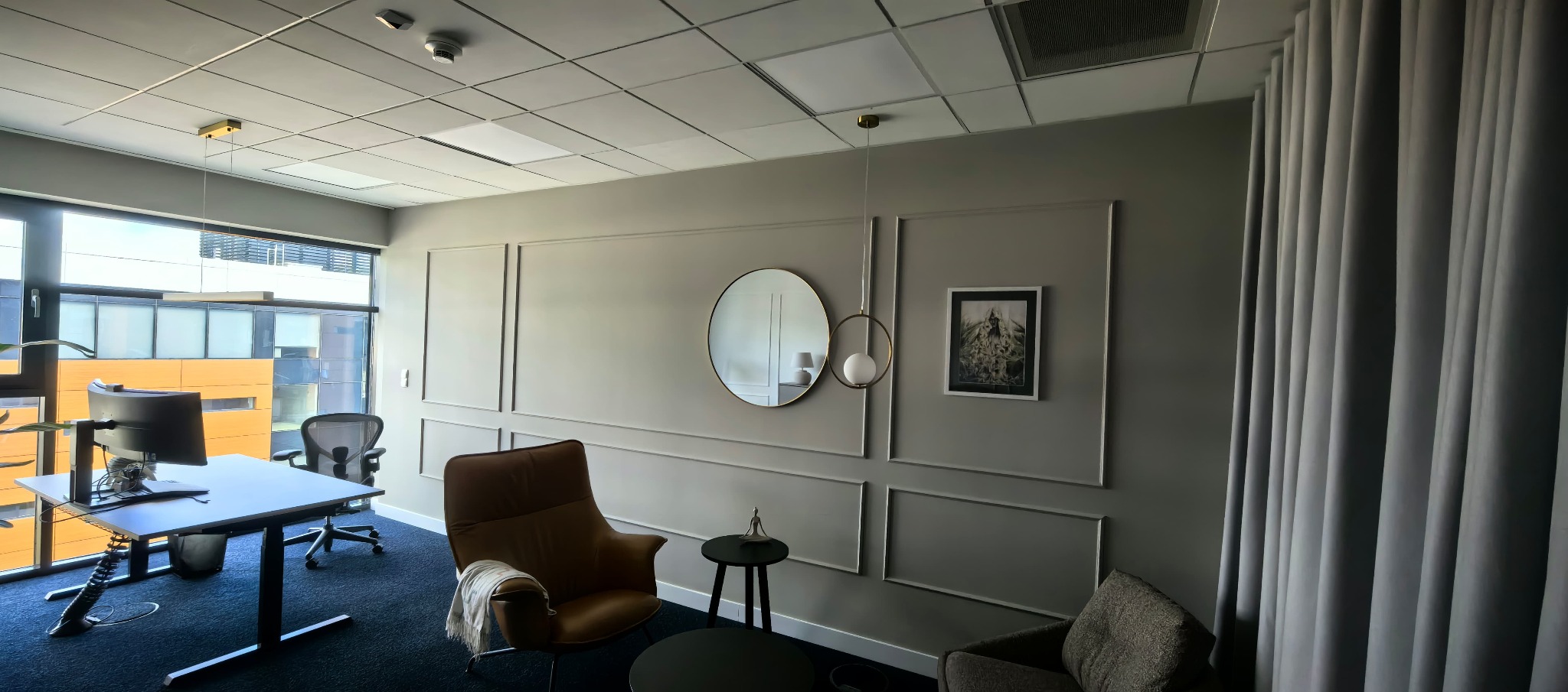 Eleganckie wykończenie wnętrza biura: szare ściany z dekoracyjnymi listwami, okrągłe lustro, designerskie lampy i wygodne fotele. Nowoczesny wystrój z dbałością o detale.