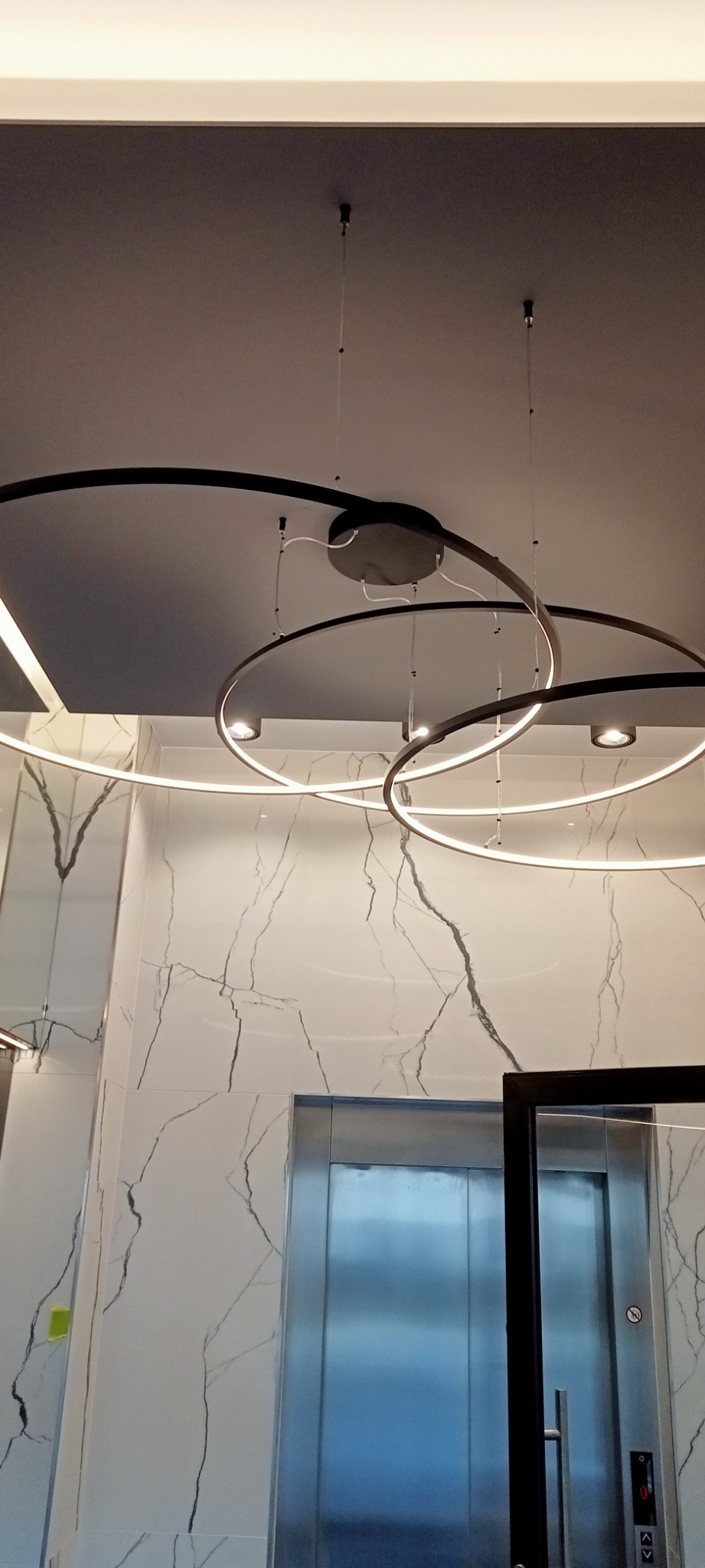 Nowoczesna lampa sufitowa z okrągłymi elementami LED, zawieszona w pomieszczeniu z marmurowymi ścianami i windą. Minimalistyczny design oświetlenia.