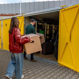 Własny Box Self Storage Warszawa Ursus - Osoby wnoszą kartony do żółtego kontenera magazynowego. Wnętrze wypełnione różnymi przedmiotami, w tym walizkami i oponami. Widoczny numer '9' na drzwiach.