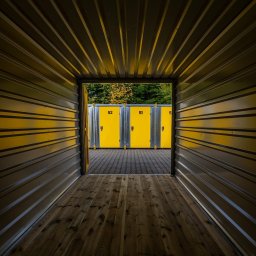 Własny Box Self Storage Warszawa Ursus - Wnętrze kontenera magazynowego z drewnianą podłogą, widok na żółte boksy z numerami 51 i 52. Perspektywa z głębi kontenera, na zewnątrz zieleń drzew.