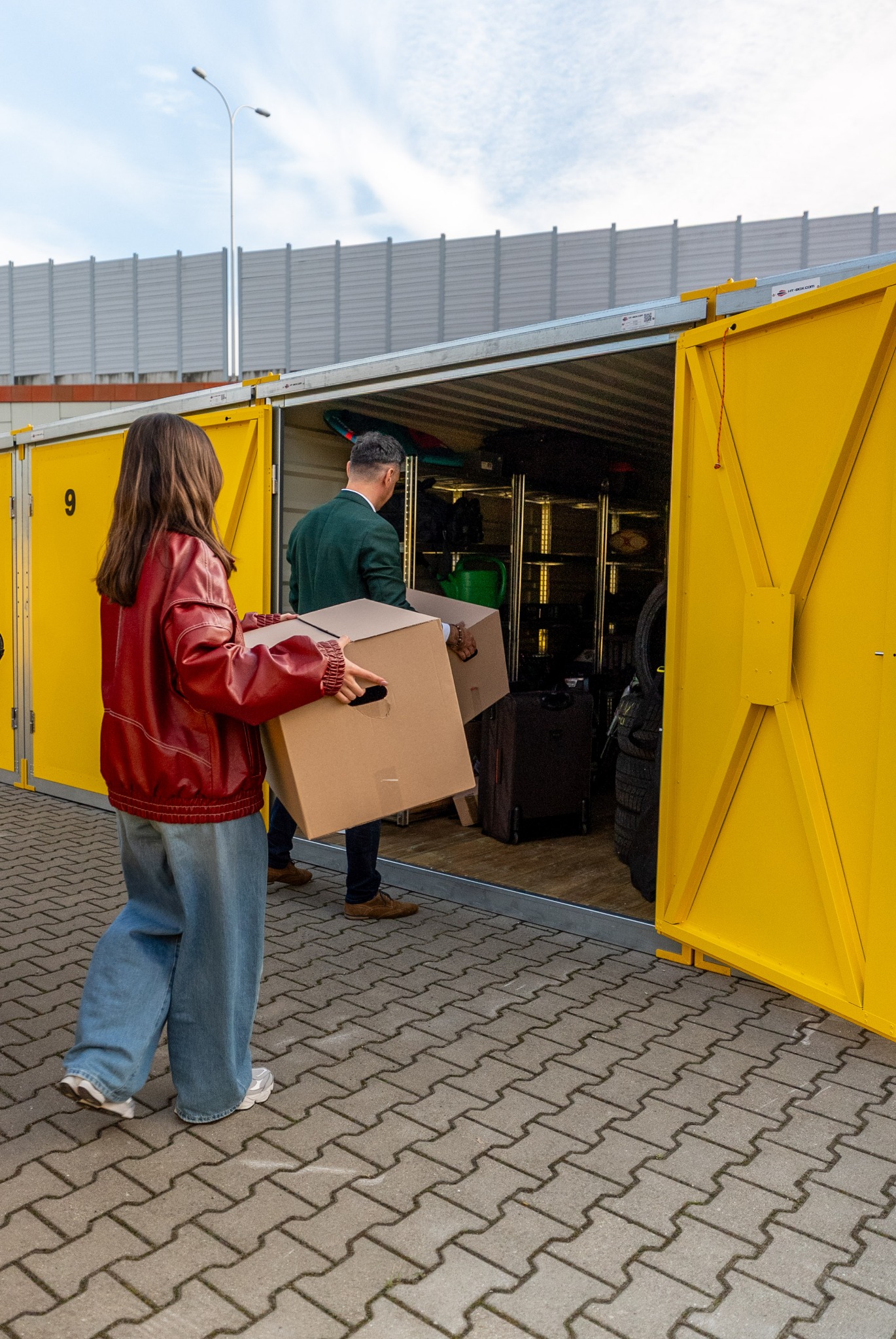 Osoby wnoszą kartony do żółtego kontenera magazynowego. Wnętrze wypełnione różnymi przedmiotami, w tym walizkami i oponami. Widoczny numer '9' na drzwiach.