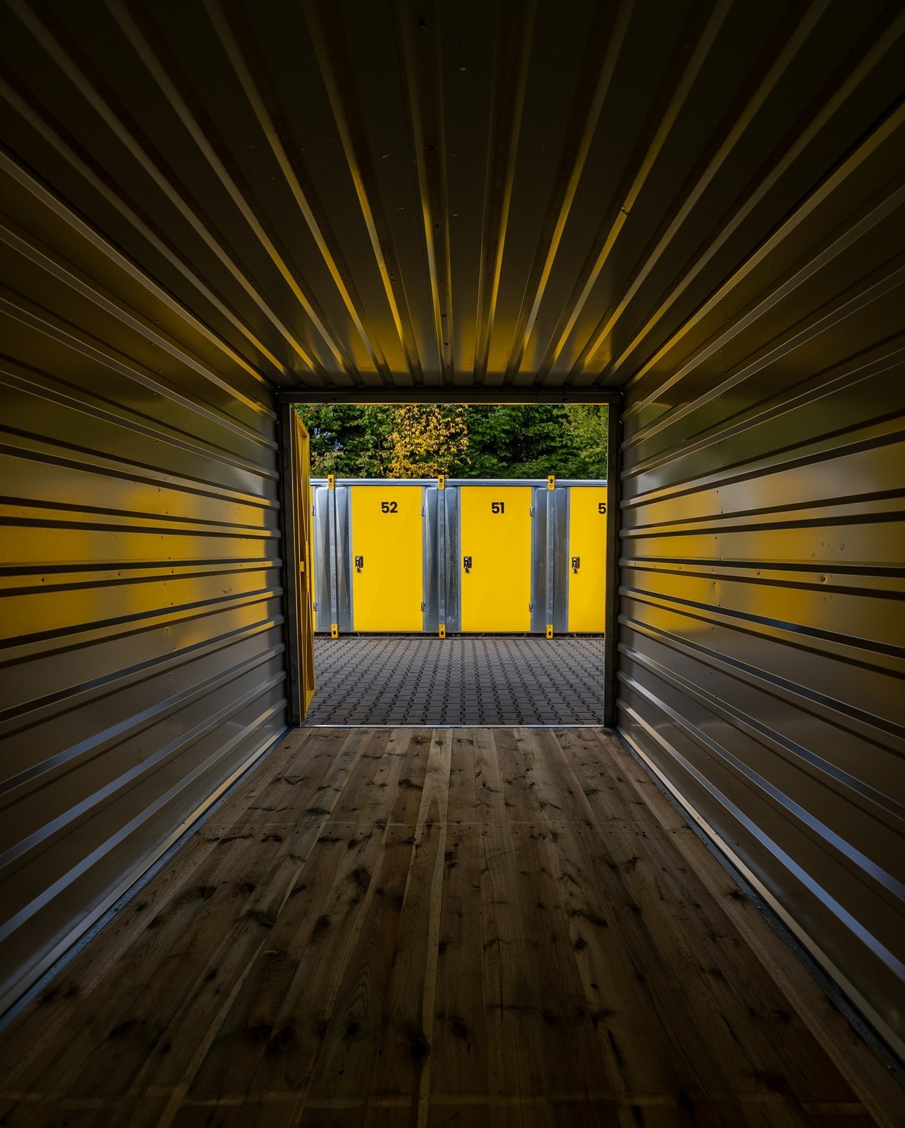Wnętrze kontenera magazynowego z drewnianą podłogą, widok na żółte boksy z numerami 51 i 52. Perspektywa z głębi kontenera, na zewnątrz zieleń drzew.