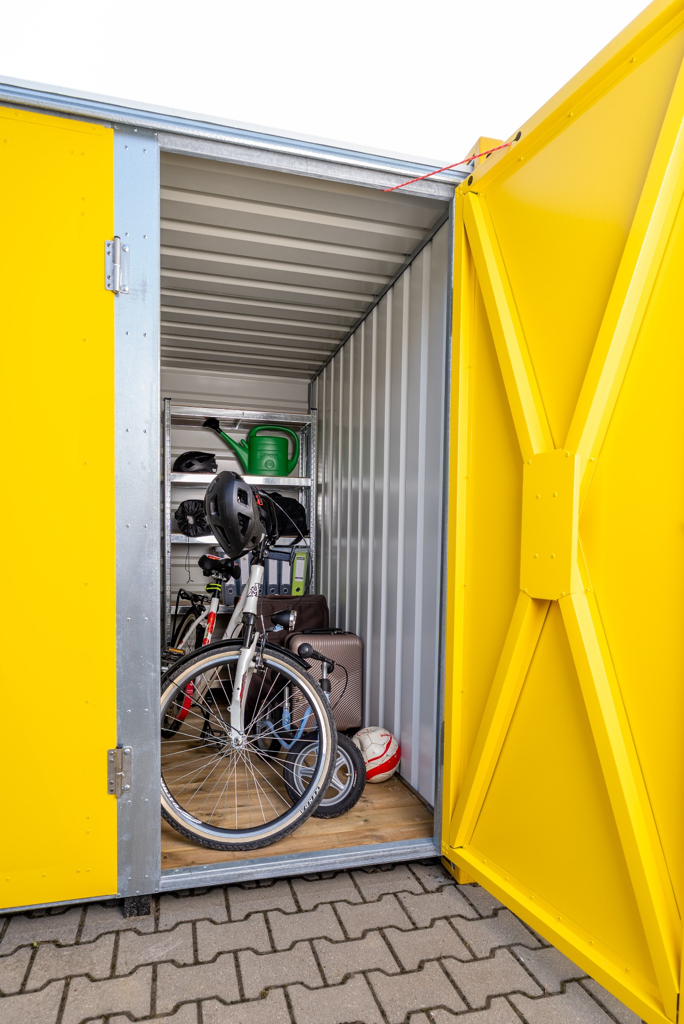 Otwarty, żółty kontener magazynowy wypełniony rowerami, walizkami i akcesoriami. Metalowe regały wewnątrz. Widok z zewnątrz na kostkę brukową.