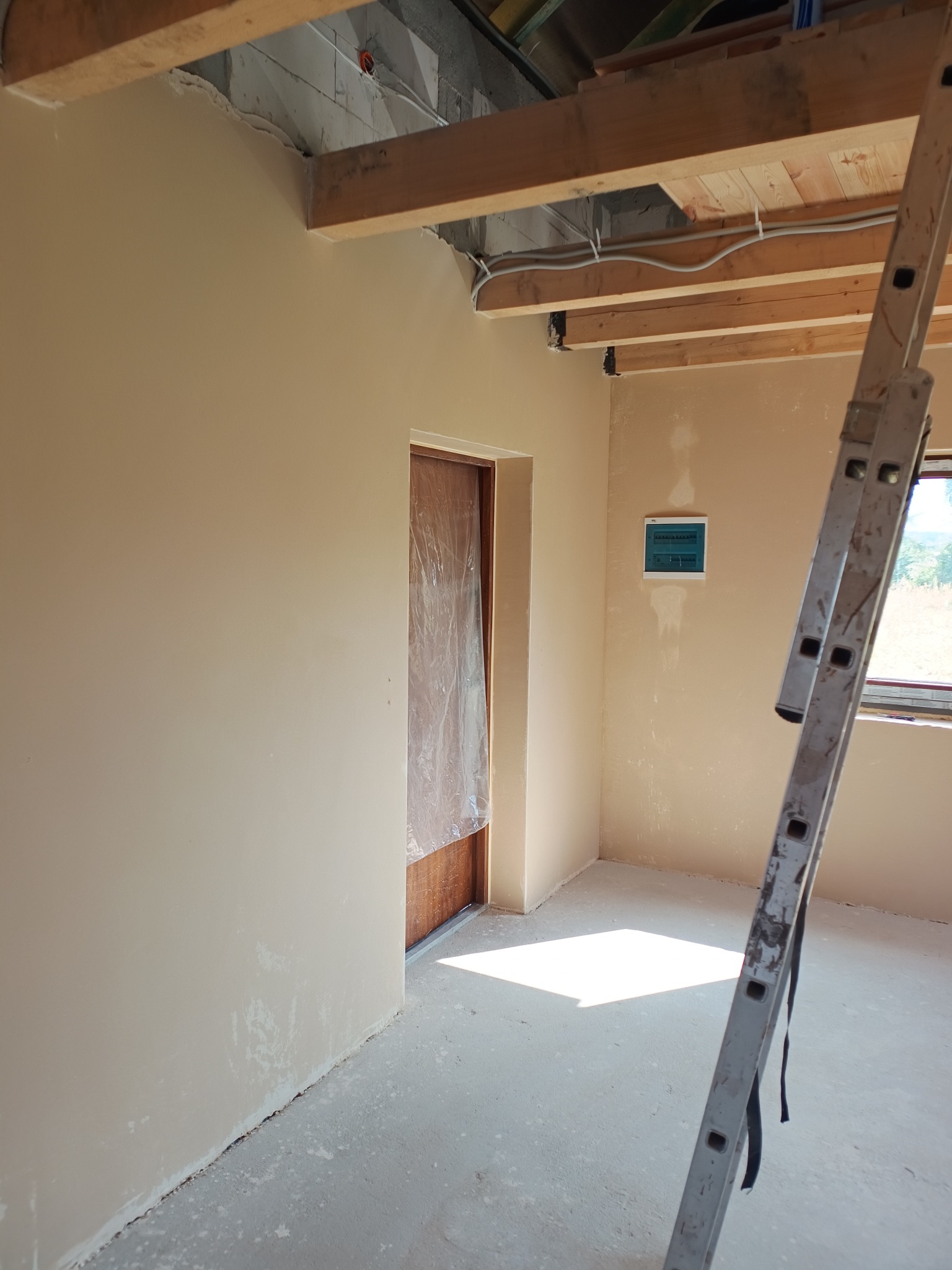 Wnętrze w trakcie prac wykończeniowych: ściany po gładzi, zabezpieczone drzwi, widoczna instalacja elektryczna pod sufitem i drabina oparta o ścianę. Jasne, naturalne światło.