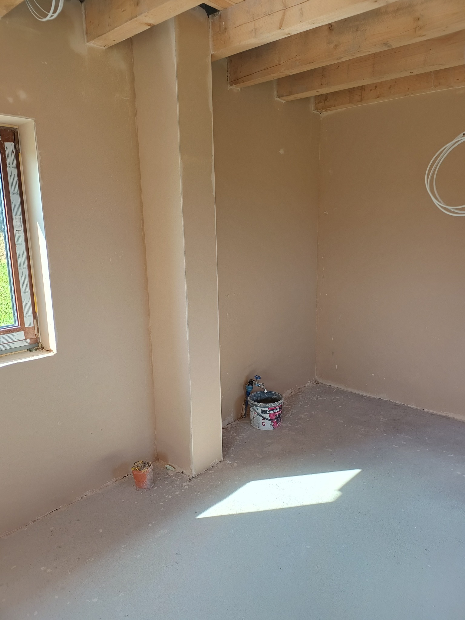 Wnętrze w trakcie prac wykończeniowych: gładkie, beżowe ściany, surowy strop drewniany, okno z widokiem na zieleń, wiadro z zaprawą i instalacja wodna przy ścianie.