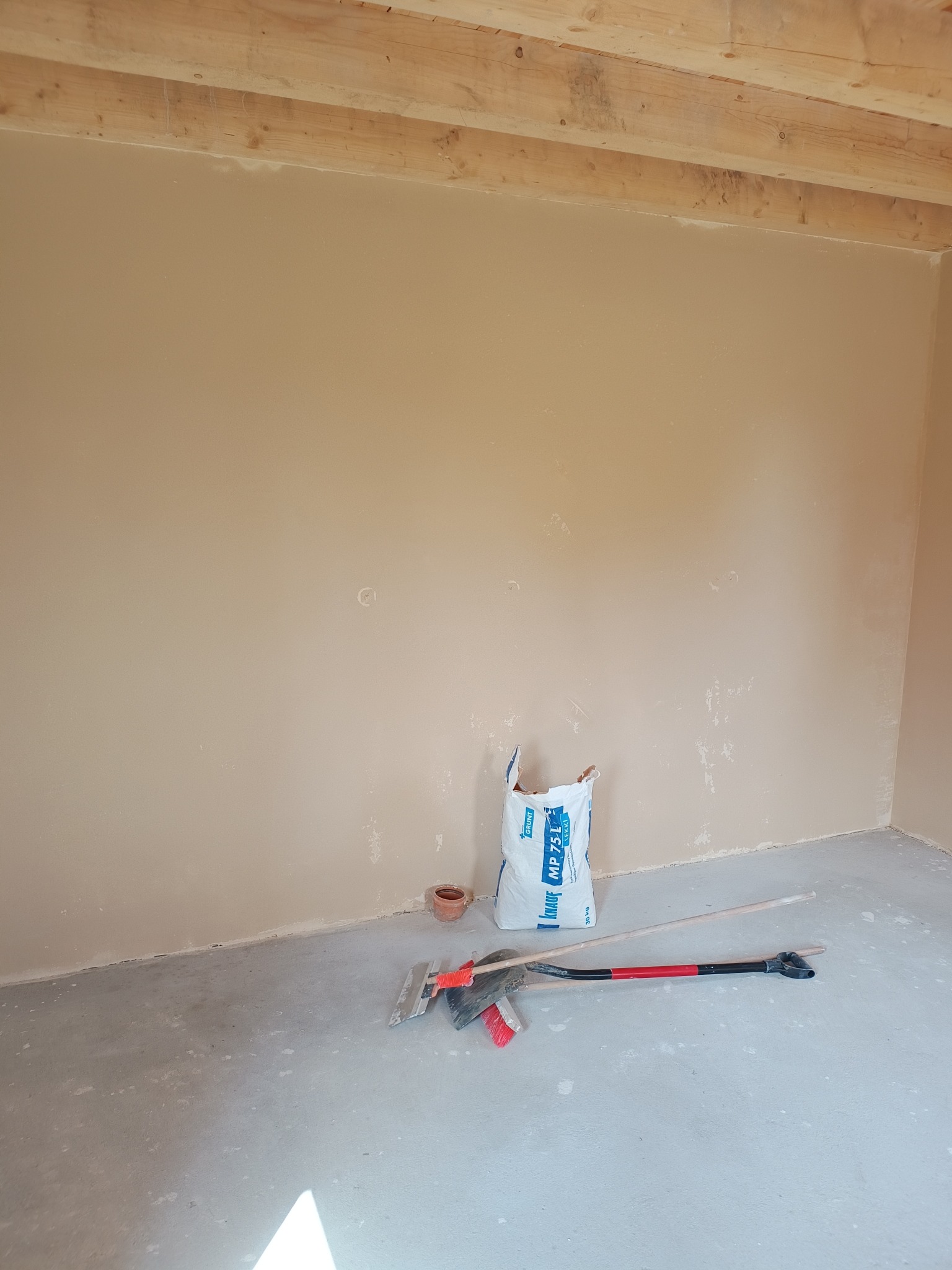 Ściana po szpachlowaniu, widoczny worek z gładzią Knauf MP 75 L, narzędzia szpachlarskie i drewniane belki stropowe. Przygotowanie do malowania.