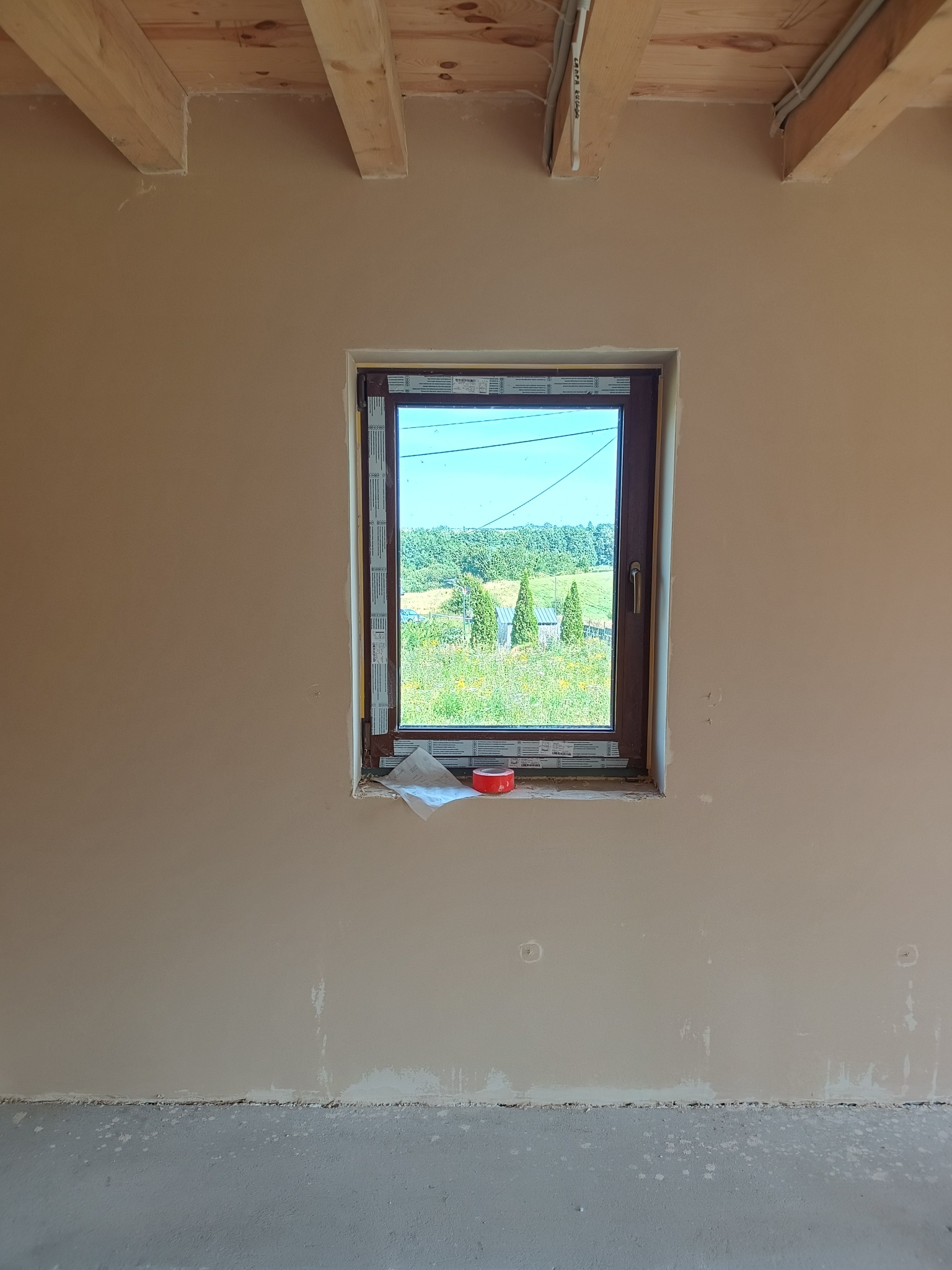 Ściana po szpachlowaniu z oknem, widoczne drewniane belki stropowe. Na parapecie czerwona taśma malarska i papier. Widok na zewnątrz przez okno.