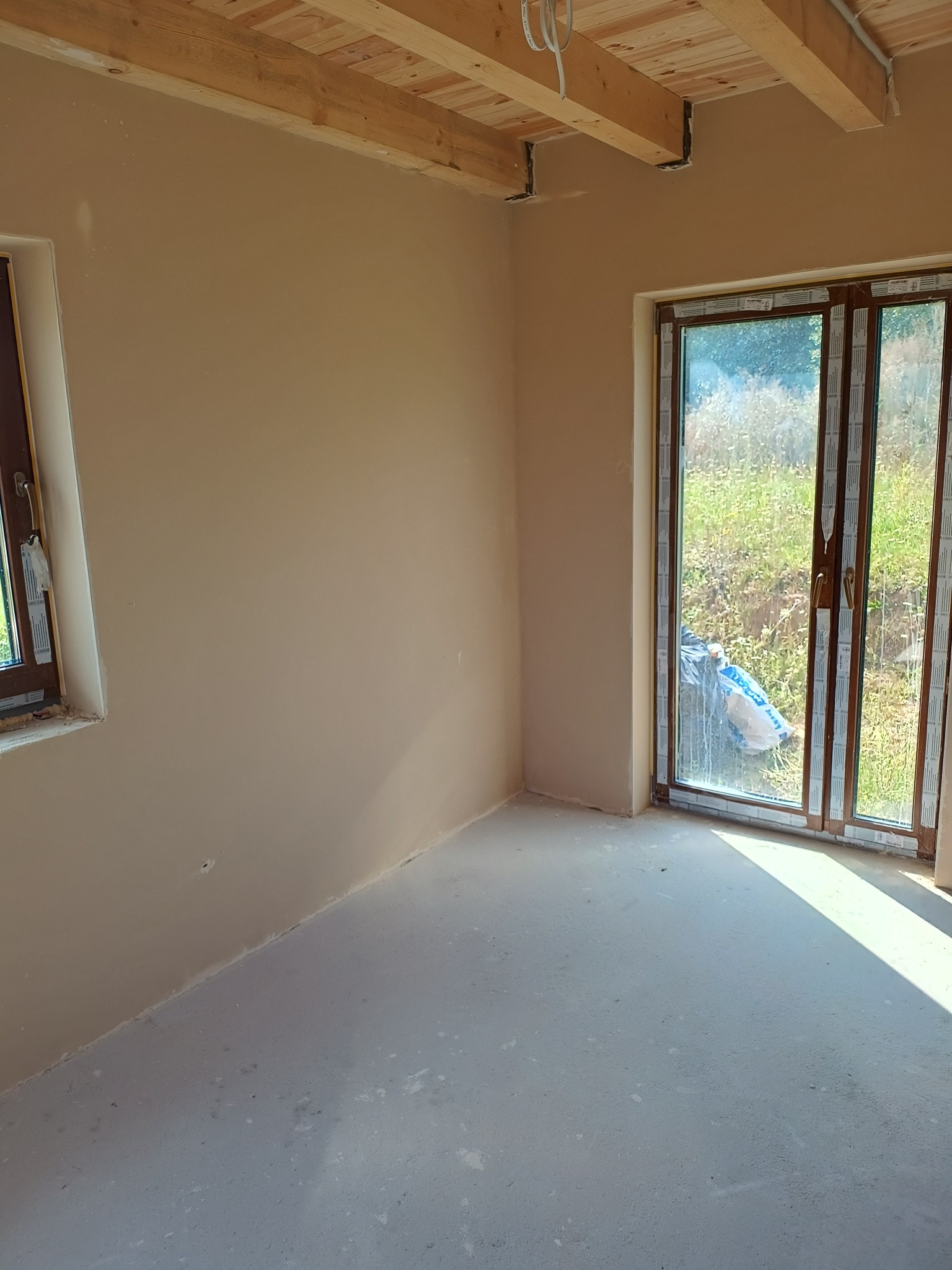 Wnętrze po szpachlowaniu: gładkie, beżowe ściany w surowym stanie, drewniany strop, okno z brązową ramą i betonowa podłoga. Widok na zewnątrz przez okno.