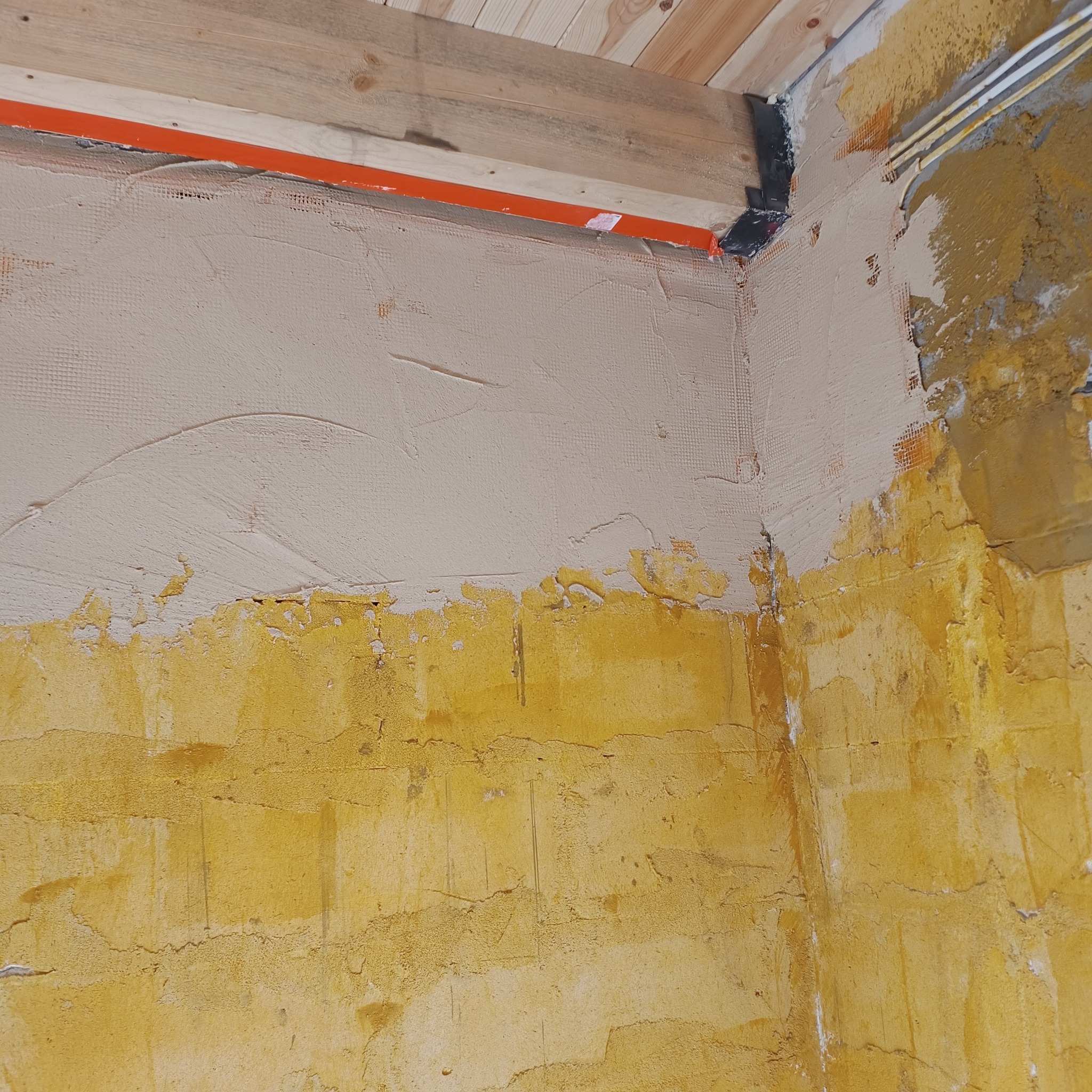 Fragment ściany w trakcie prac wykończeniowych: dolna część w kolorze żółtym, górna pokryta siatką i świeżo nałożoną, jasną warstwą gładzi. Widoczne elementy drewniane sufitu.
