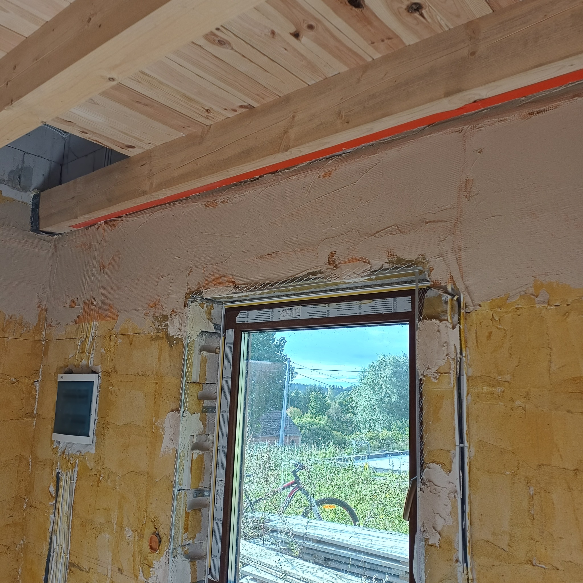 Wnętrze w trakcie szpachlowania: ściany z nałożoną siatką i częściowo wygładzone, widoczne drewniane belki stropowe i okno z widokiem na zieleń.