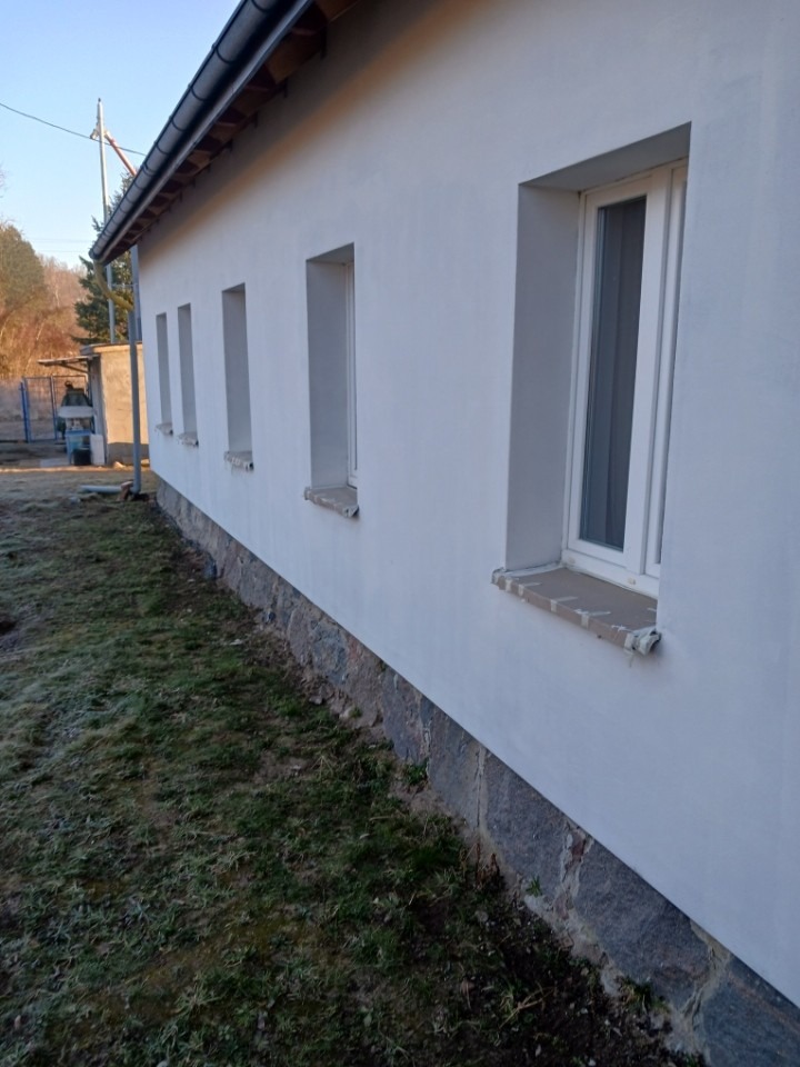 Boczny widok budynku po renowacji elewacji, z widocznymi oknami i kamienną podmurówką. Świeżo pomalowana, jasna ściana kontrastuje z zielenią trawy.