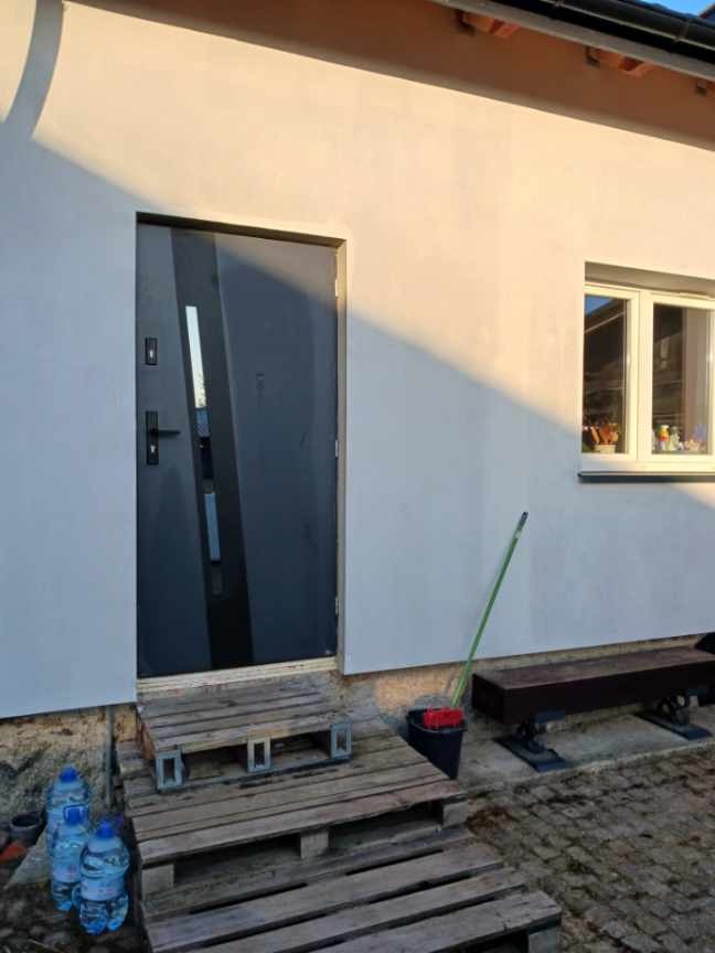 Biała elewacja domu jednorodzinnego z szarą stolarką drzwiową i oknem. Widoczne palety jako tymczasowe schody i narzędzia malarskie.