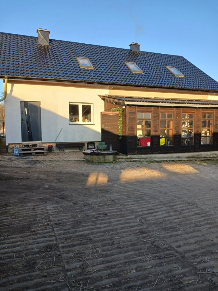 Dom jednorodzinny z jasną elewacją, ciemnym dachem z oknami połaciowymi i drewnianą werandą. Widoczne elementy wykończeniowe i otoczenie budynku.