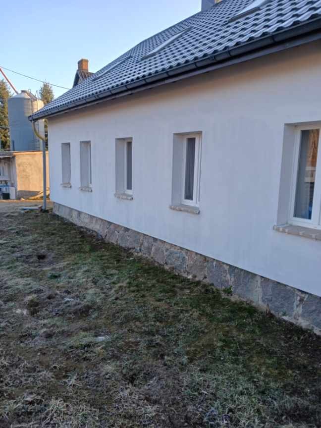 Boczny widok domu z jasną elewacją i ciemnym dachem. Widoczne okna z białymi ramami i kamienny cokół. W tle drzewa i budynki gospodarcze.