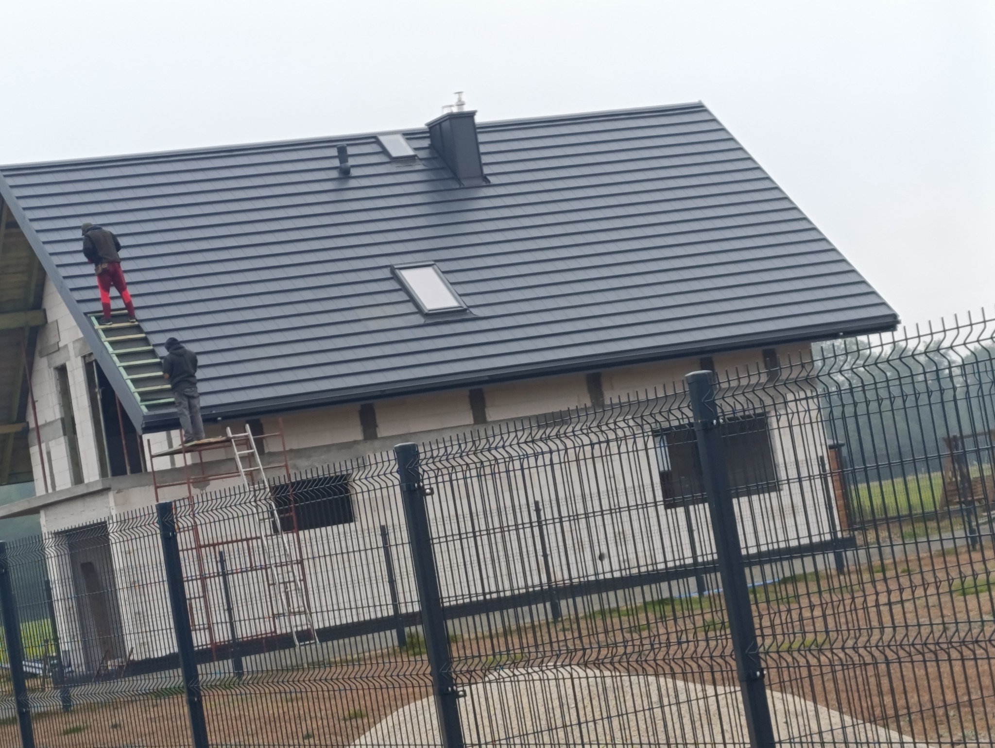 Montaż czarnej blachodachówki na domu w stanie surowym otwartym. Dwóch dekarzy na dachu, widoczne rusztowania i ogrodzenie posesji. Dach z oknami dachowymi i kominem.