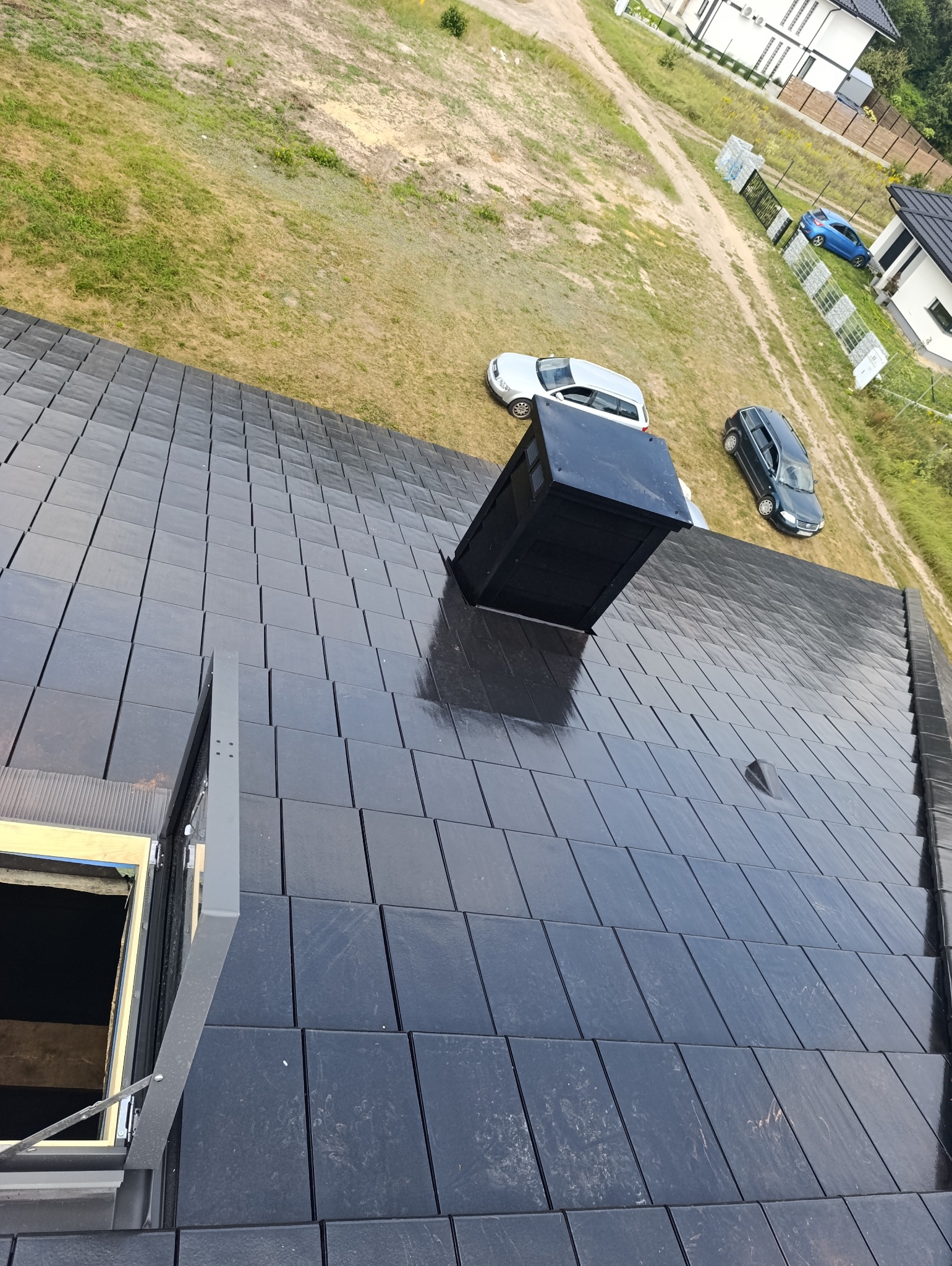 Błyszczący, grafitowy dach z dachówki ceramicznej, z kominem i oknem dachowym. W tle trawnik, domy i zaparkowane auta. Ujęcie z góry.