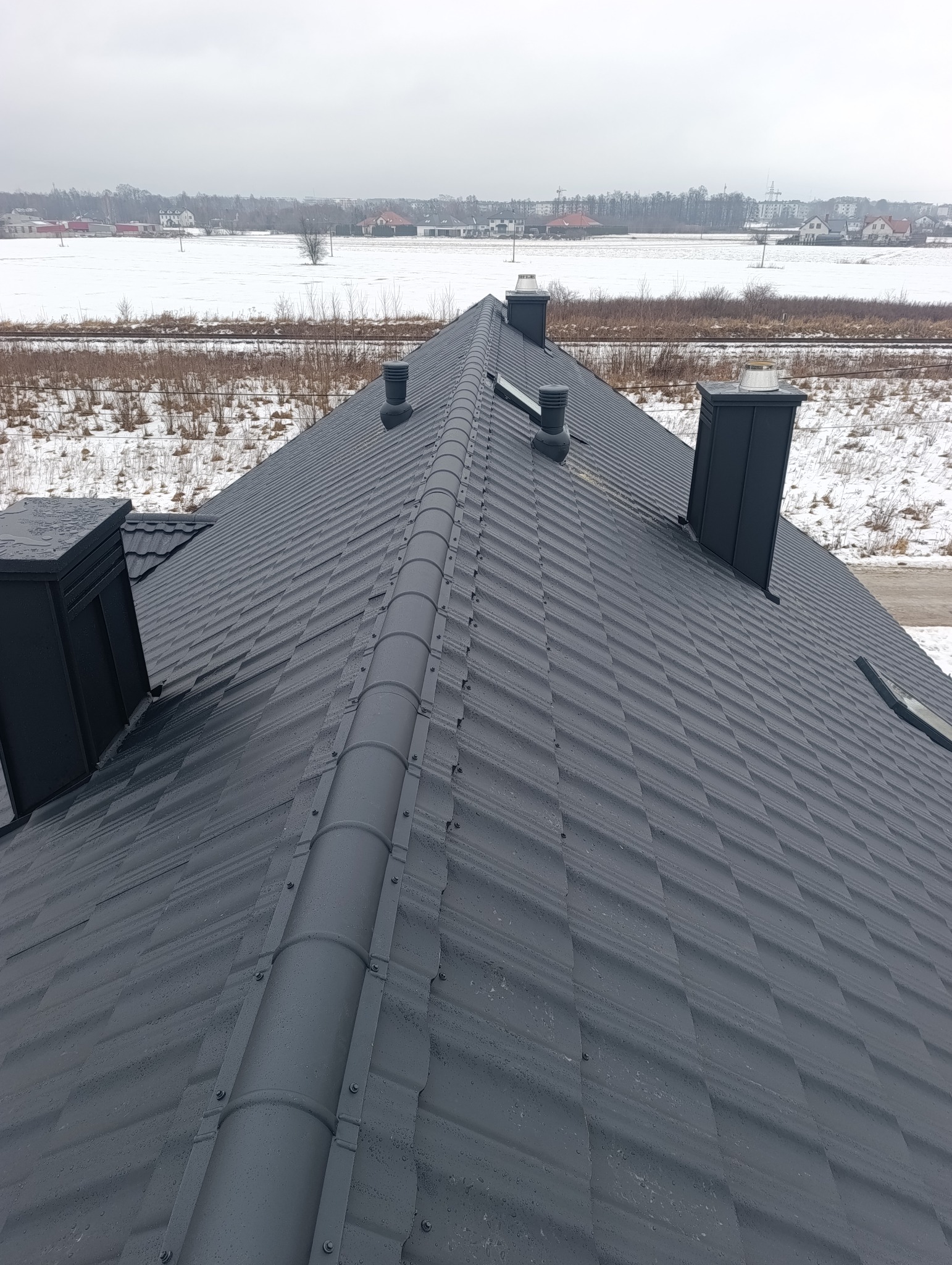 Grafitowy dach z blachy trapezowej z kominami i odpowietrznikami na tle zimowego krajobrazu. Widoczne krople wody na powierzchni dachu.