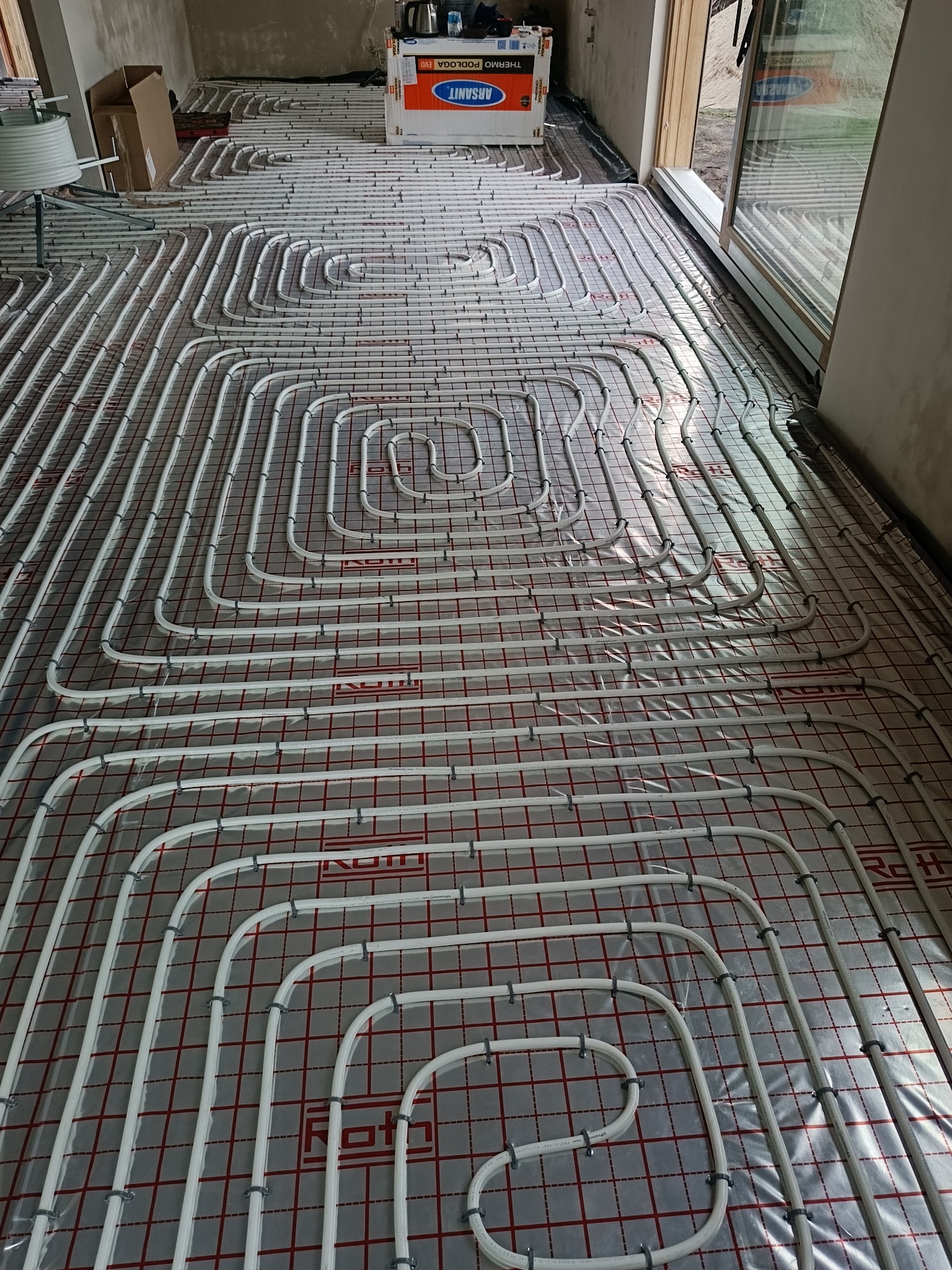Białe rury ogrzewania podłogowego ułożone na folii z czerwoną kratką, widok z góry, przed wylaniem posadzki. Na górze kartonowe opakowanie materiałów budowlanych.