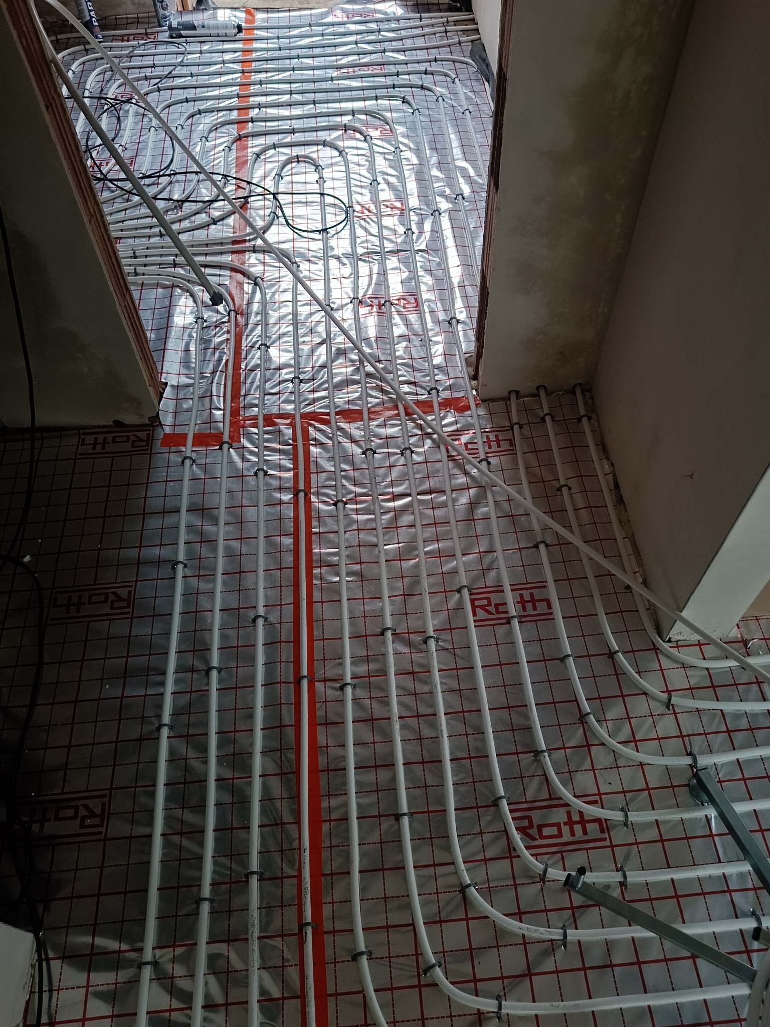 Białe rury ogrzewania podłogowego Roth, ułożone na folii z czerwoną siatką, widoczne uchwyty mocujące, surowy stan wykończenia ścian i podłogi.