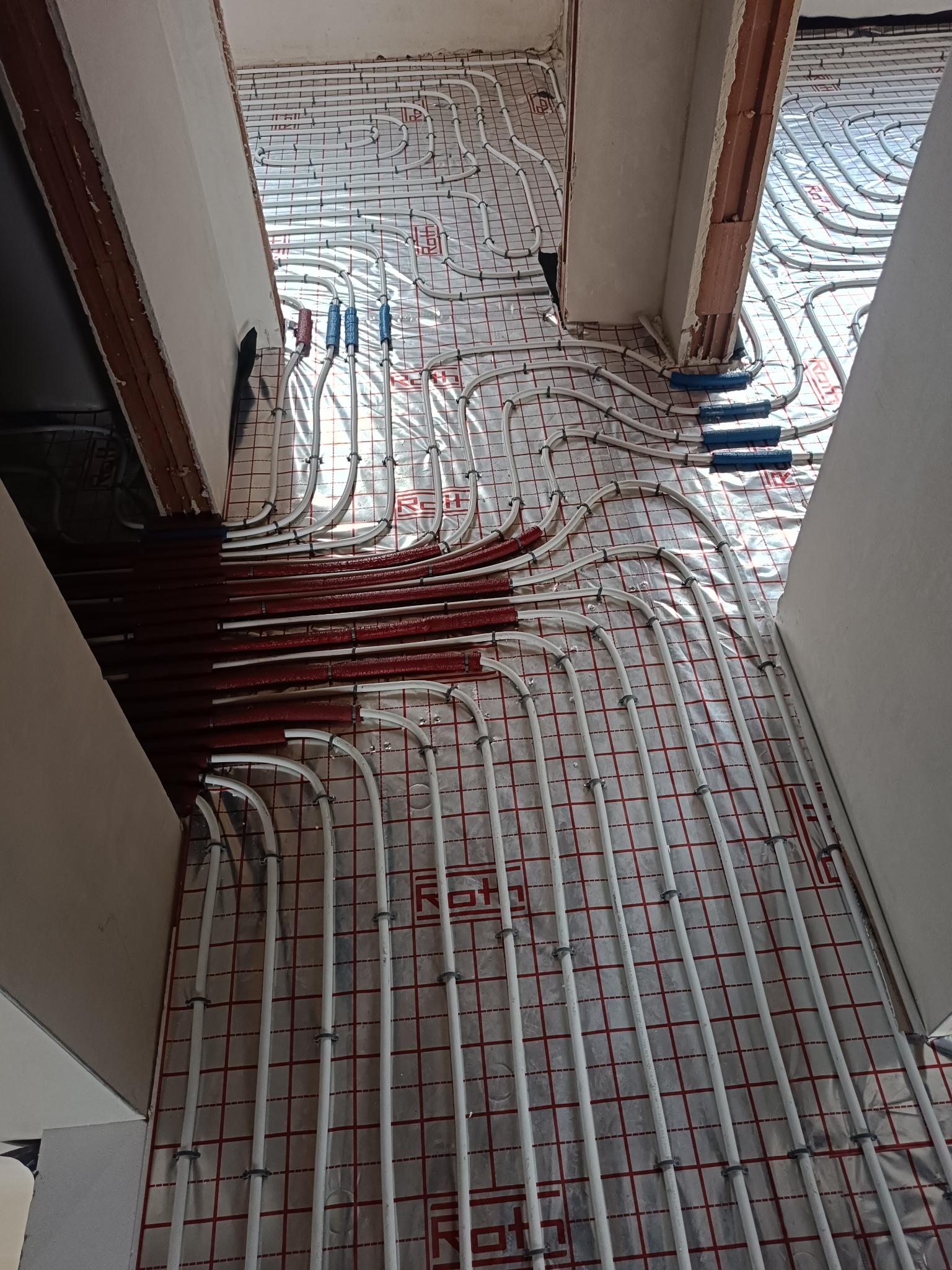 Białe rury ogrzewania podłogowego ułożone na folii z czerwoną siatką, widoczne fragmenty ścian i przejść między pomieszczeniami. Część rur pomalowana na czerwono.