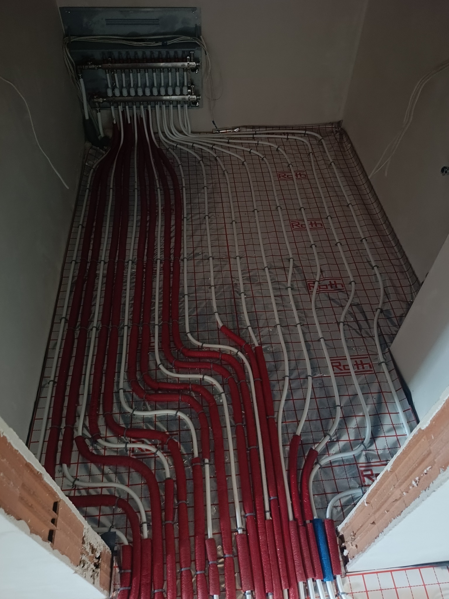 Rozłożona instalacja ogrzewania podłogowego z czerwonymi i białymi rurami na izolacji z siatką, widok z góry, z kolektorem na ścianie.