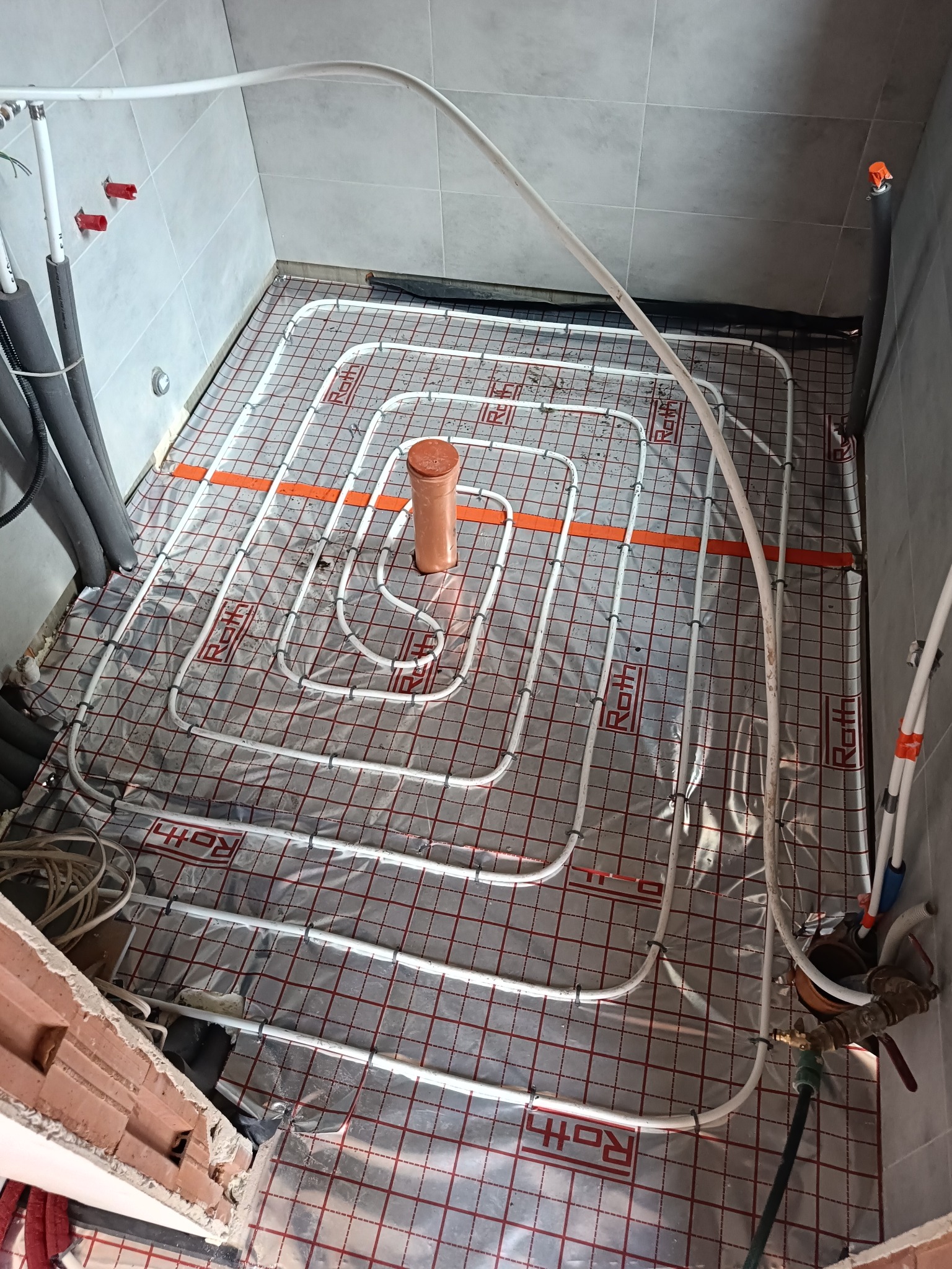 Białe rury ogrzewania podłogowego ułożone spiralnie na folii z czerwoną kratką w łazience z szarymi płytkami. Widoczne szare rury doprowadzające.