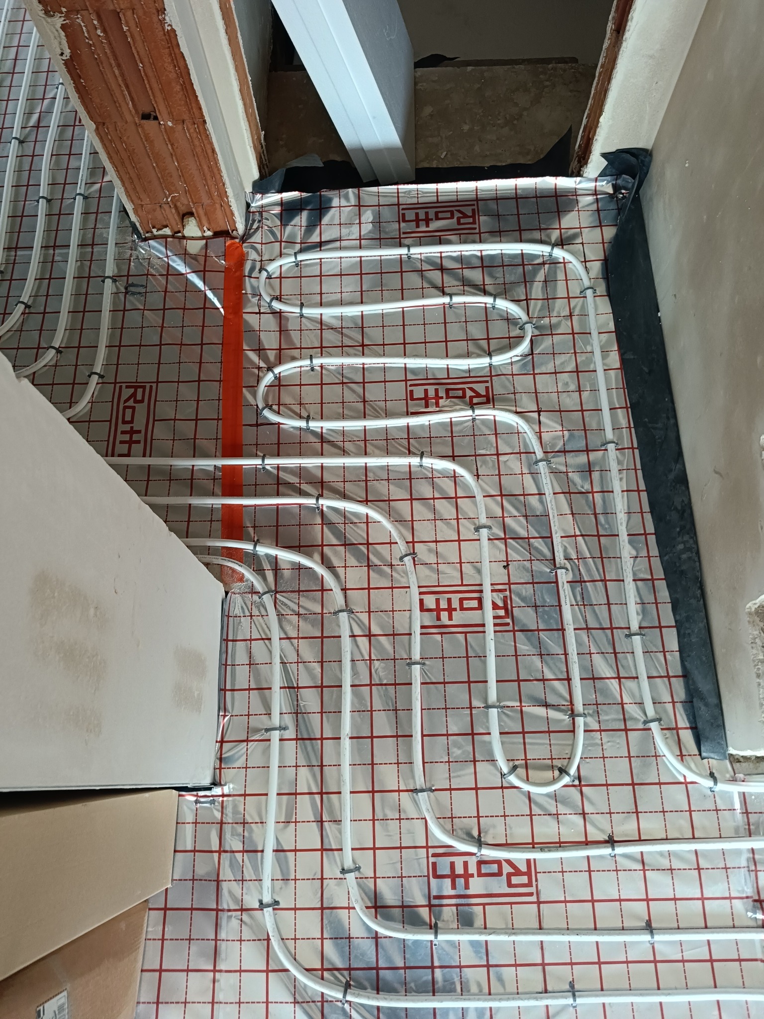 Białe rury ogrzewania podłogowego Roth, zamocowane na folii z czerwoną siatką, widoczne w trakcie instalacji w pomieszczeniu z surowymi ścianami.